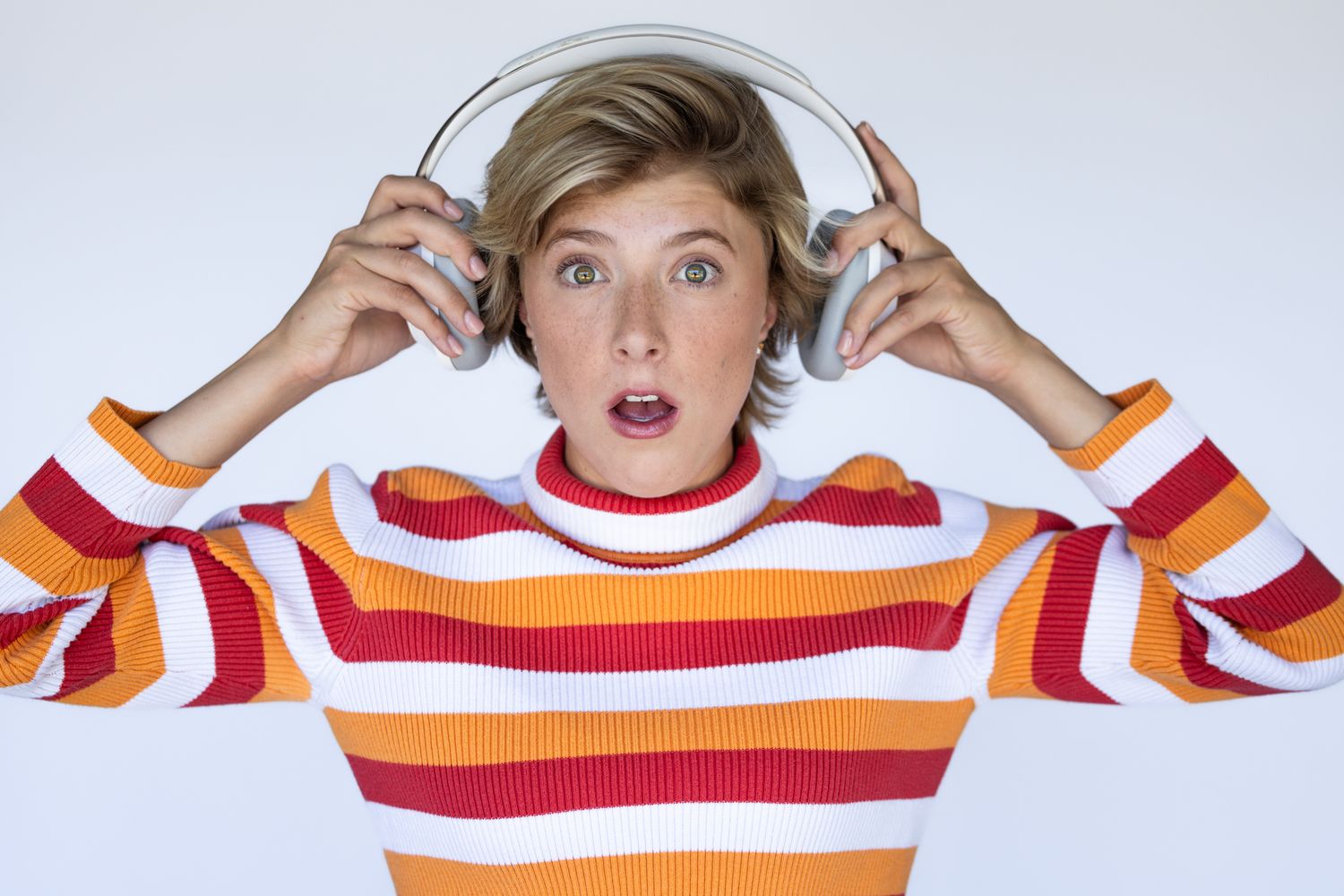 Someone in a striped red and orange sweater puts on headphones with a surprised expression against a white background.