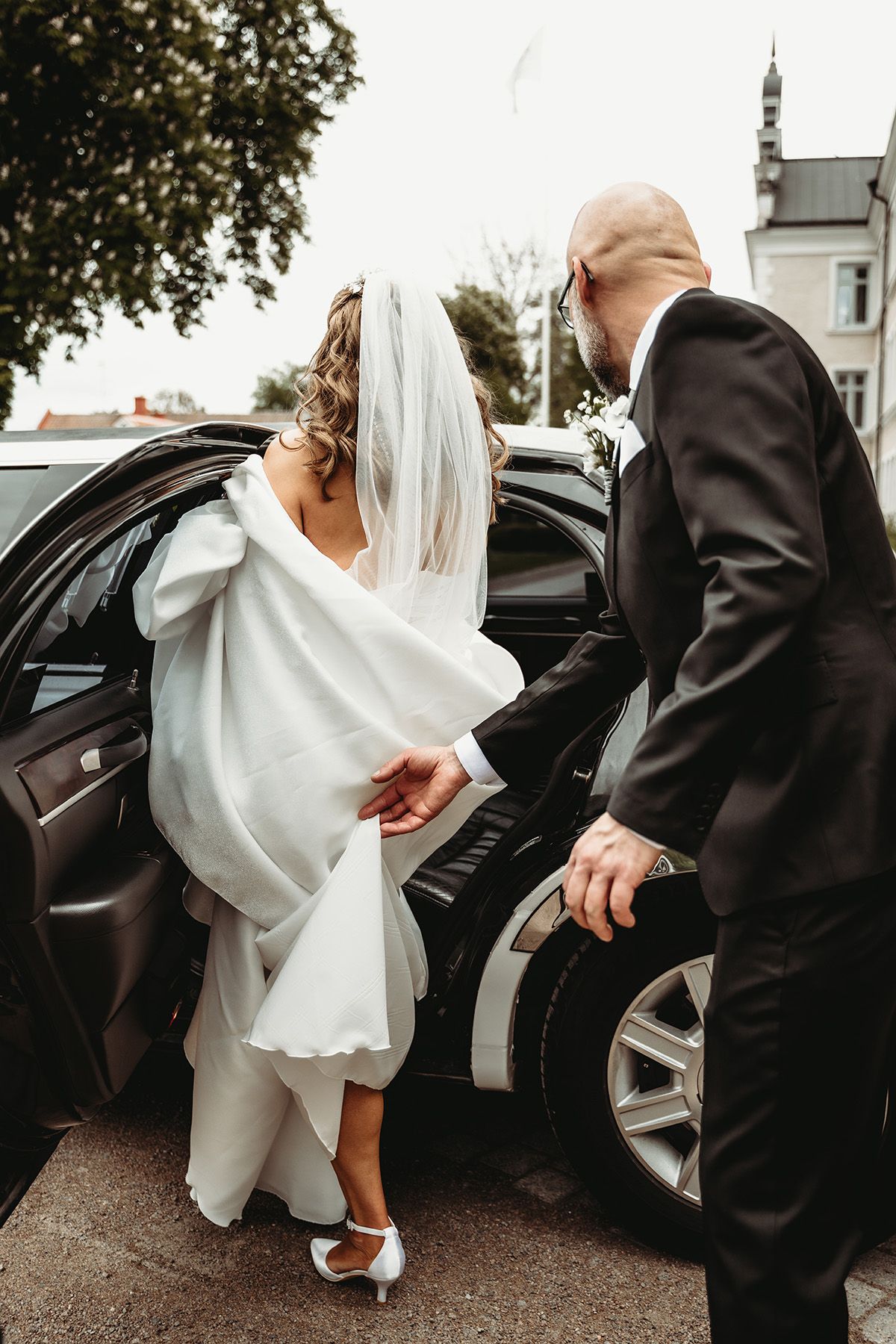 Brud i vacker vit klänning och slöja kliver in i en limousine, brudgummen hjälper henne hålla upp släpet. Bröllopsfoto i Vadstena.