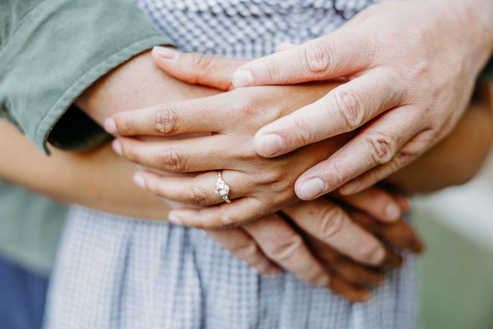 Hands with engagement ring are tenderly clasped together against a blue and sage green fabric background.