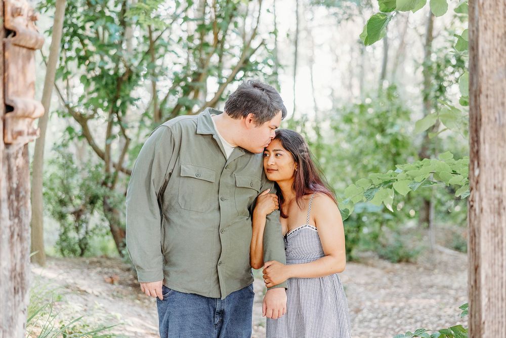 A romantic couple shares an intimate moment while walking together on a wooded trail in natural lighting.