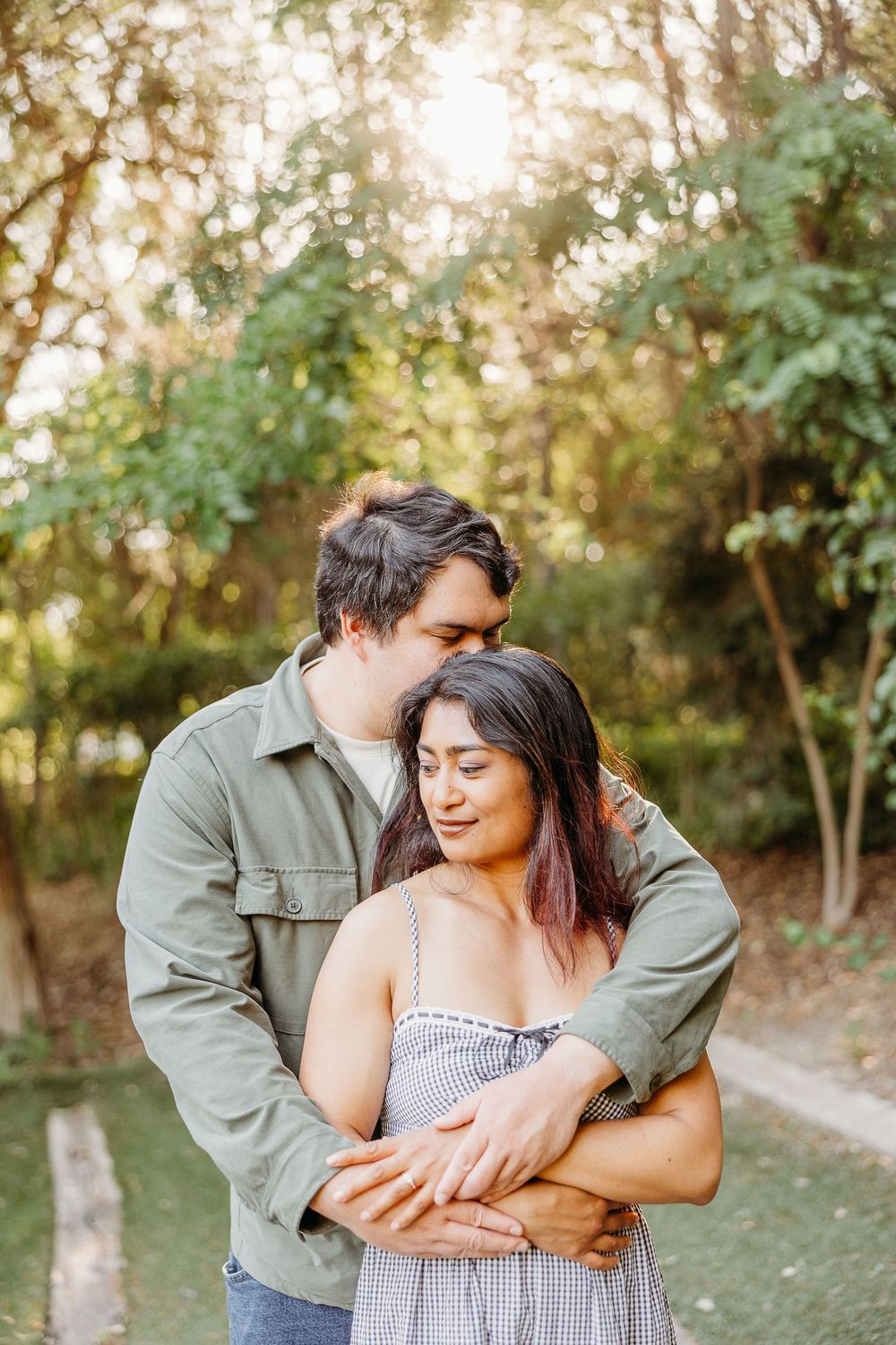 A couple shares an intimate embrace in a sunlit outdoor setting with trees and greenery in the background.
