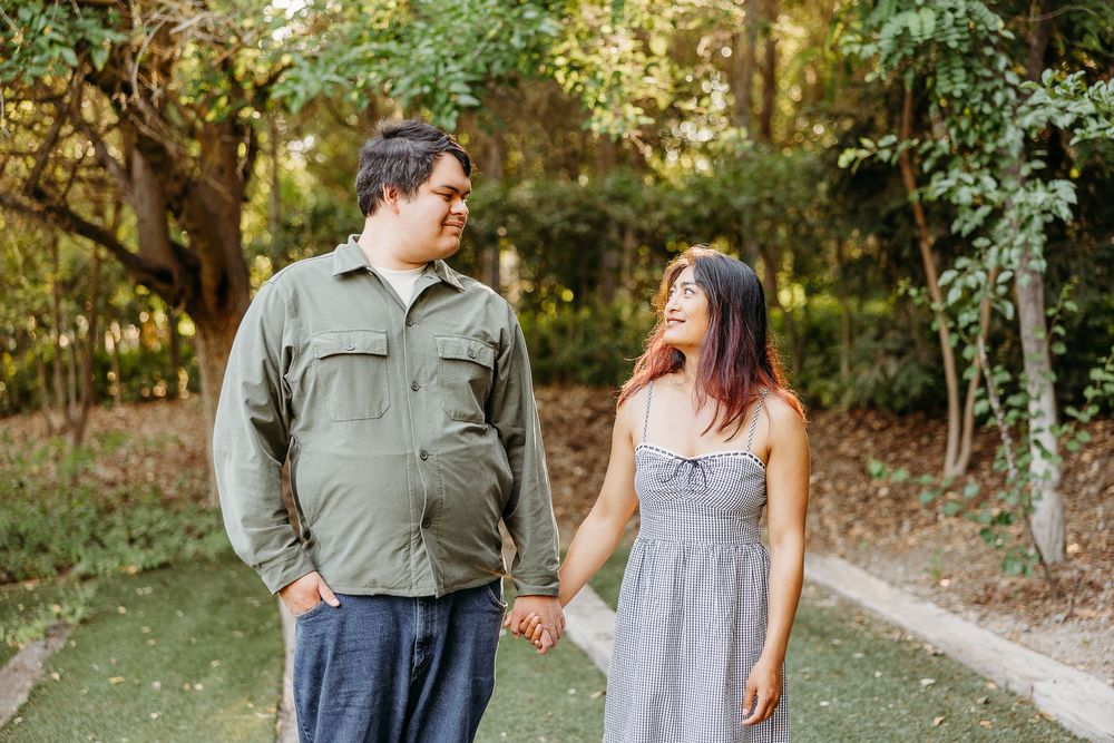 A couple holds hands while walking on a path surrounded by lush green trees and foliage.