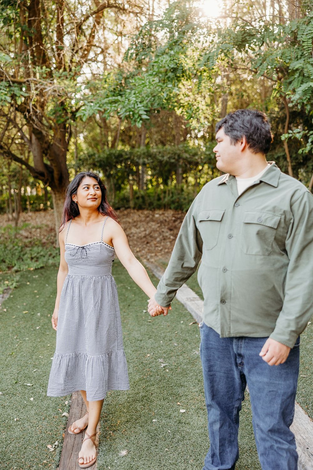 A couple holding hands while walking together in a garden setting, with her wearing a gray dress and him in casual attire.