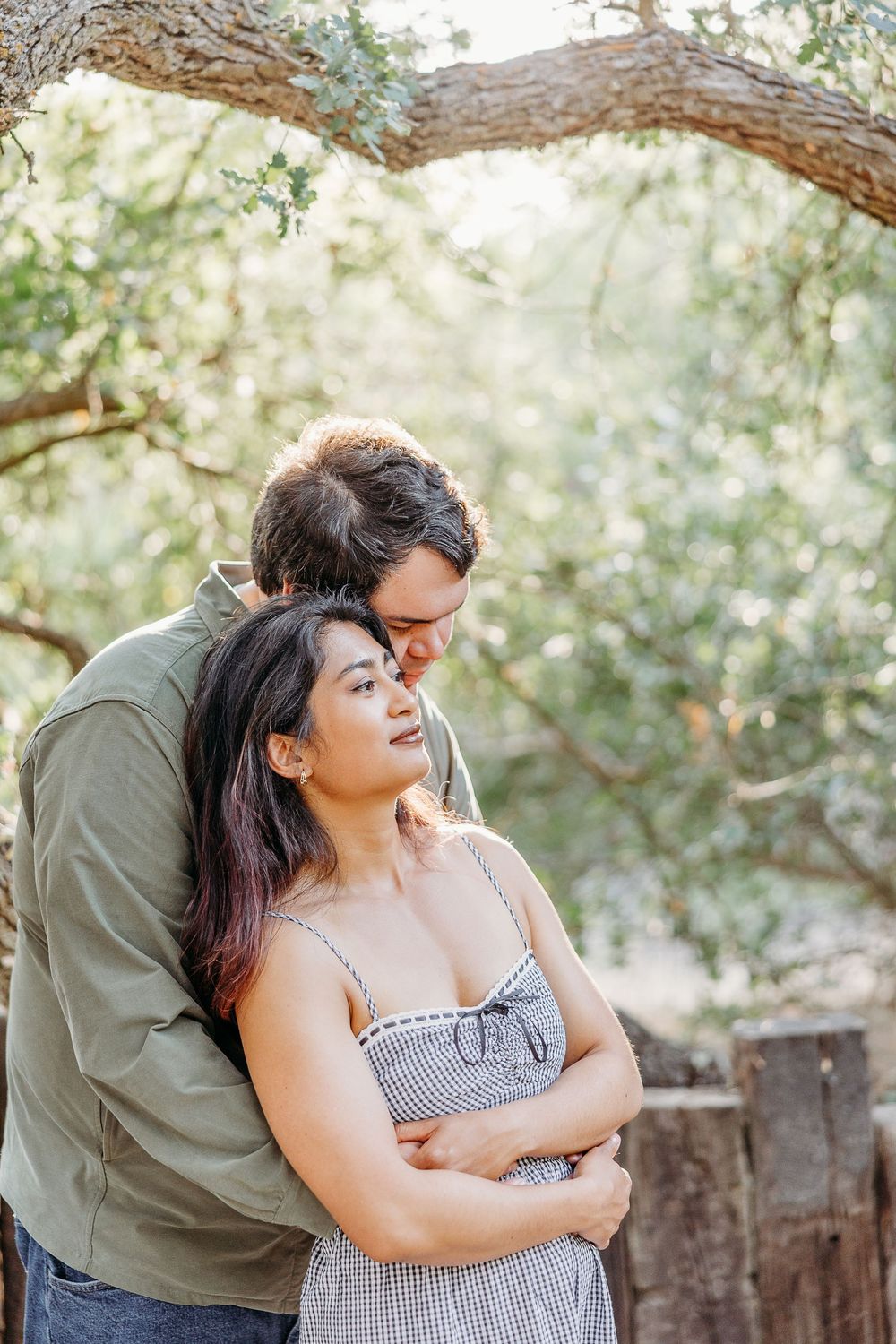 A couple embraces under a tree branch in a sun-dappled outdoor setting on a peaceful afternoon.