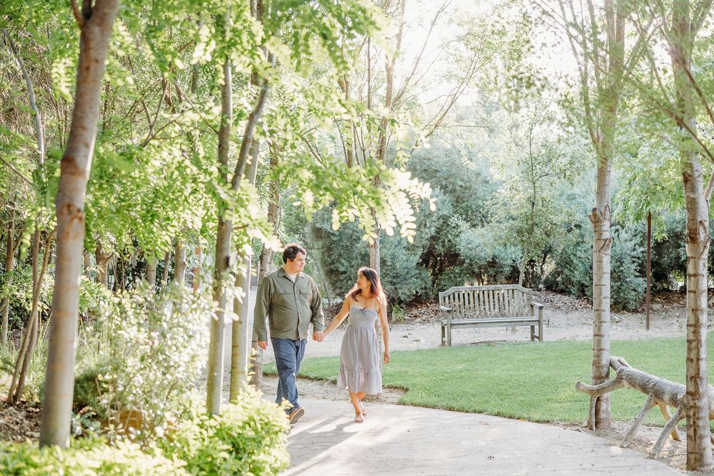 Two people stroll along a winding garden path surrounded by lush trees and a decorative bench in the background.