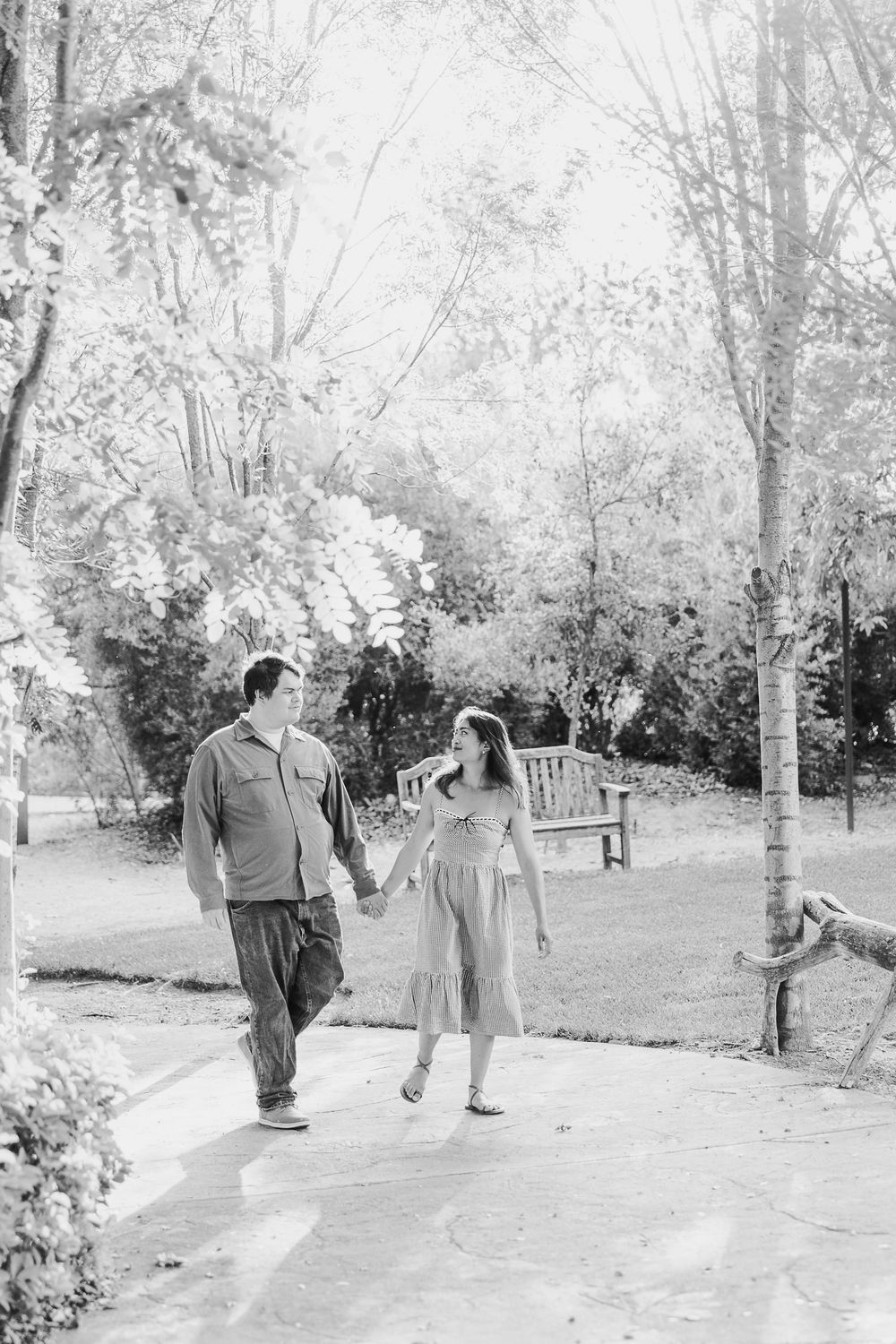 Two figures stroll together along a serene garden path surrounded by trees and natural scenery in black and white.