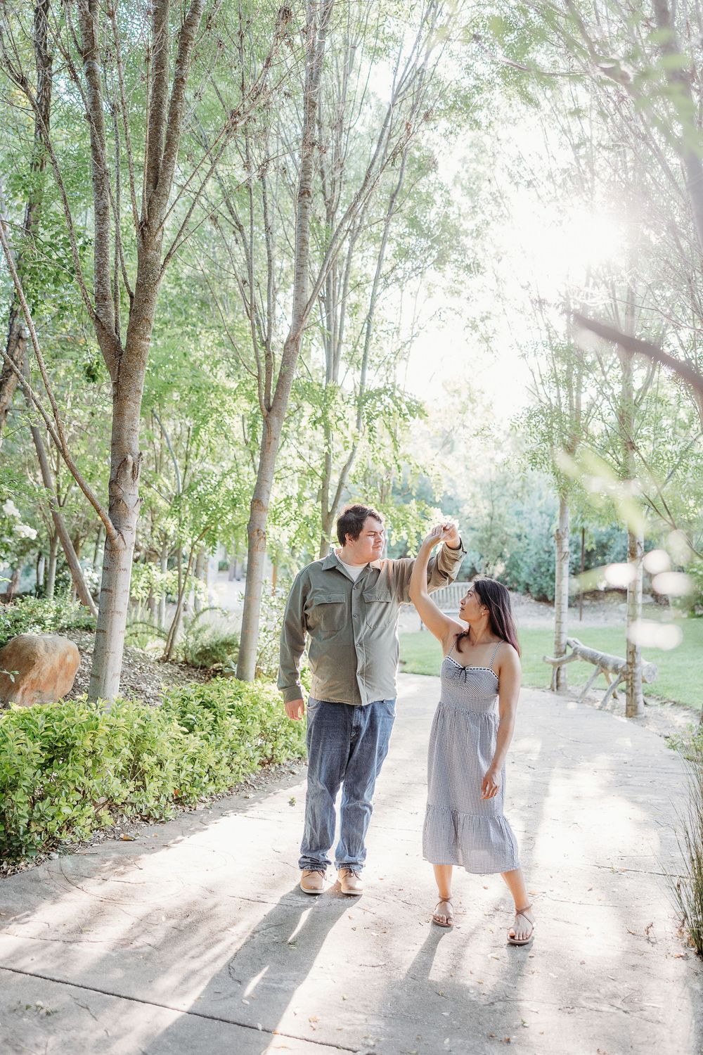 A couple walking together along a sunlit tree-lined pathway in a garden setting on a bright sunny day.