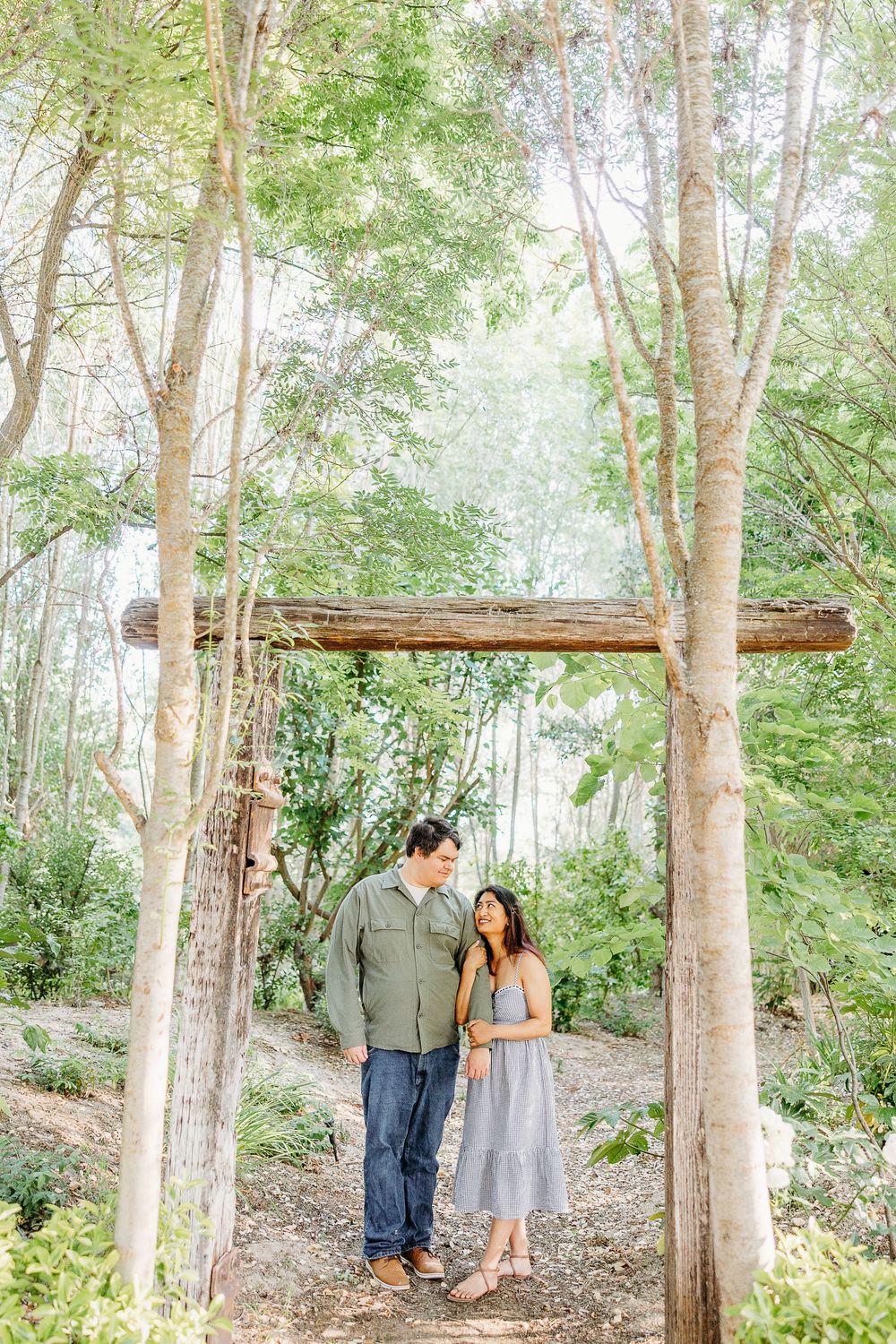 A couple sharing a romantic moment under a rustic wooden arch in a dreamy forest setting.
