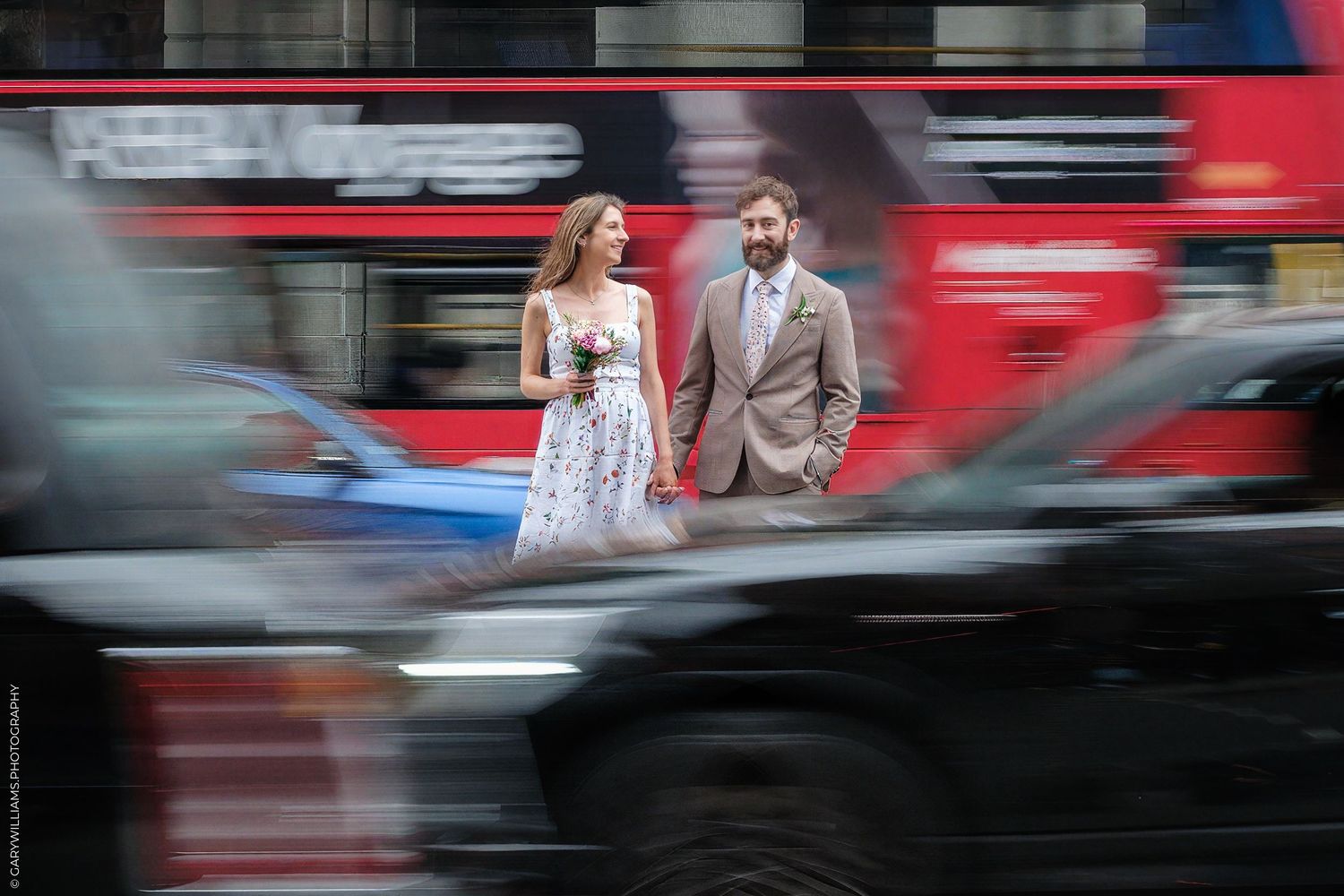 Wedding couple poses for romantic photos in front of motion-blurred red London buses on city street.