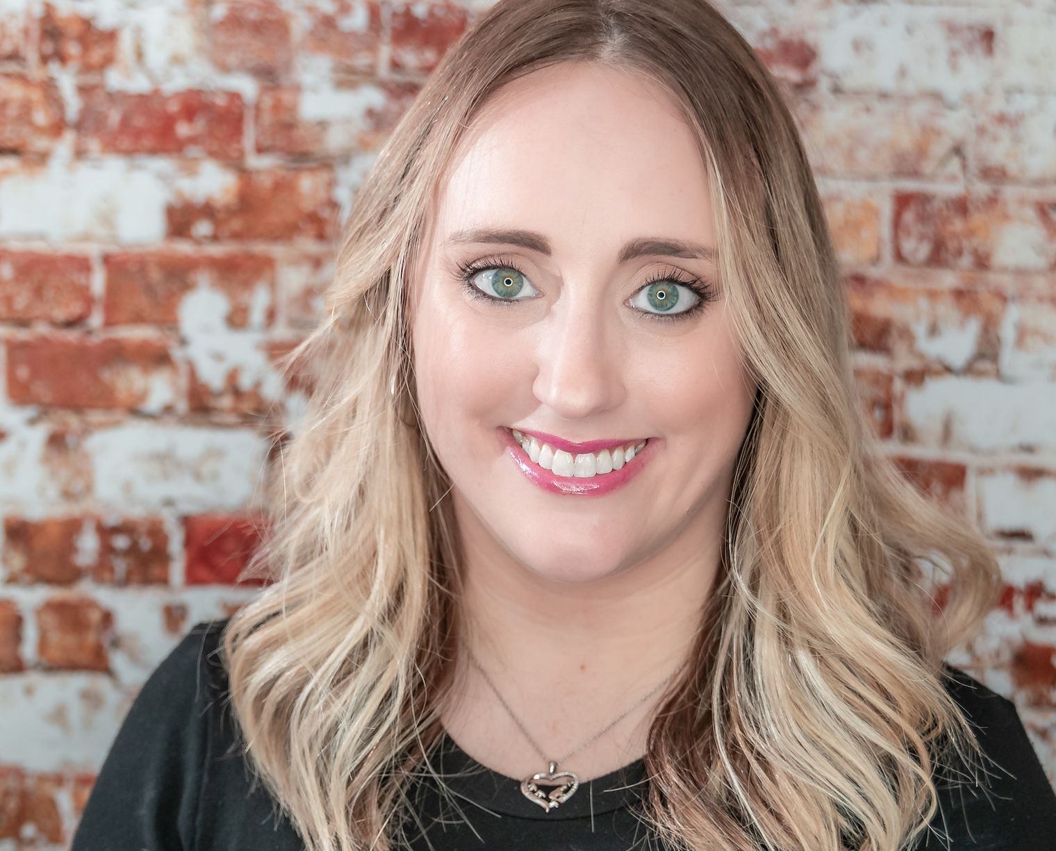Woman with blonde wavy hair and bright smile poses against rustic brick wall backdrop.