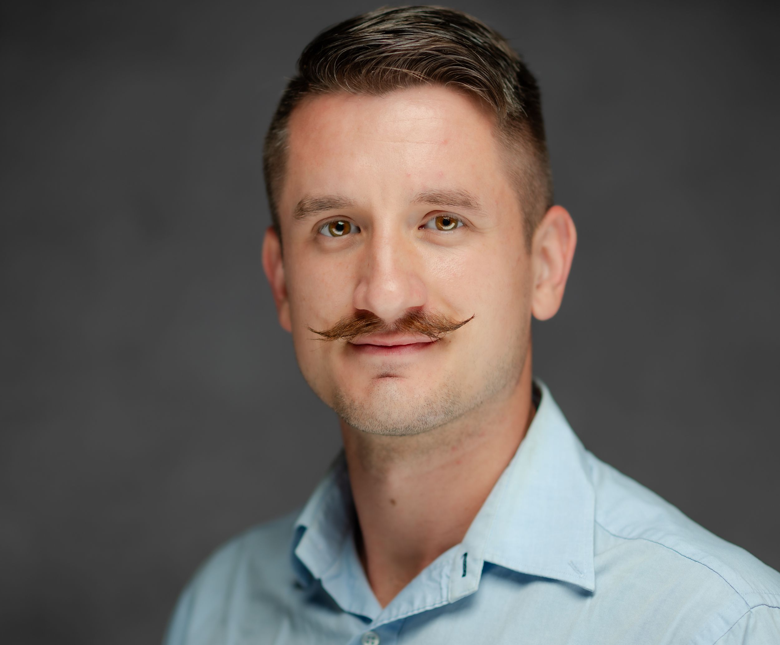 Professional headshot of someone wearing a light blue button-down shirt with a well-groomed handlebar mustache.