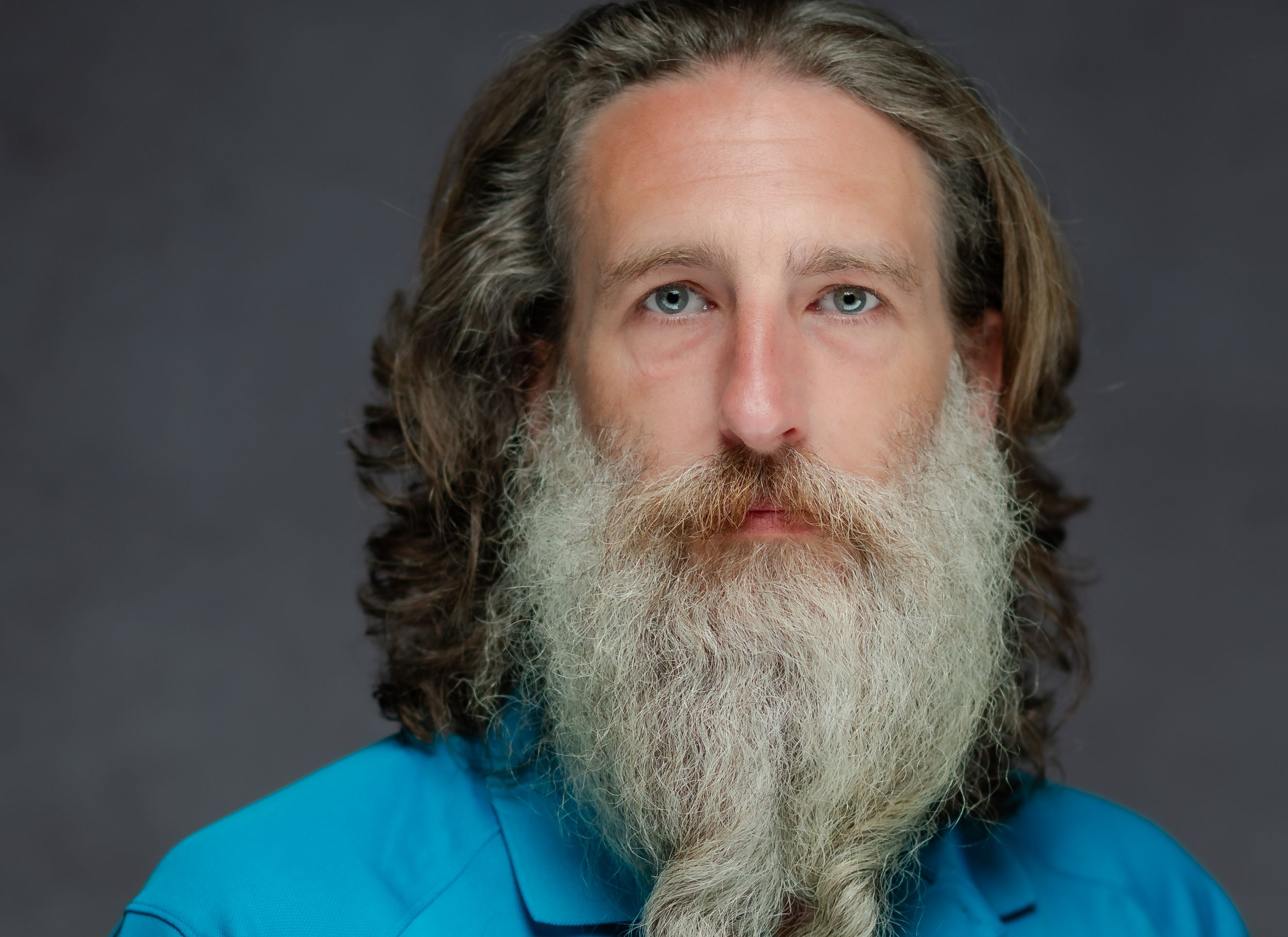 Person with long gray beard wearing bright blue shirt poses against dark studio background.