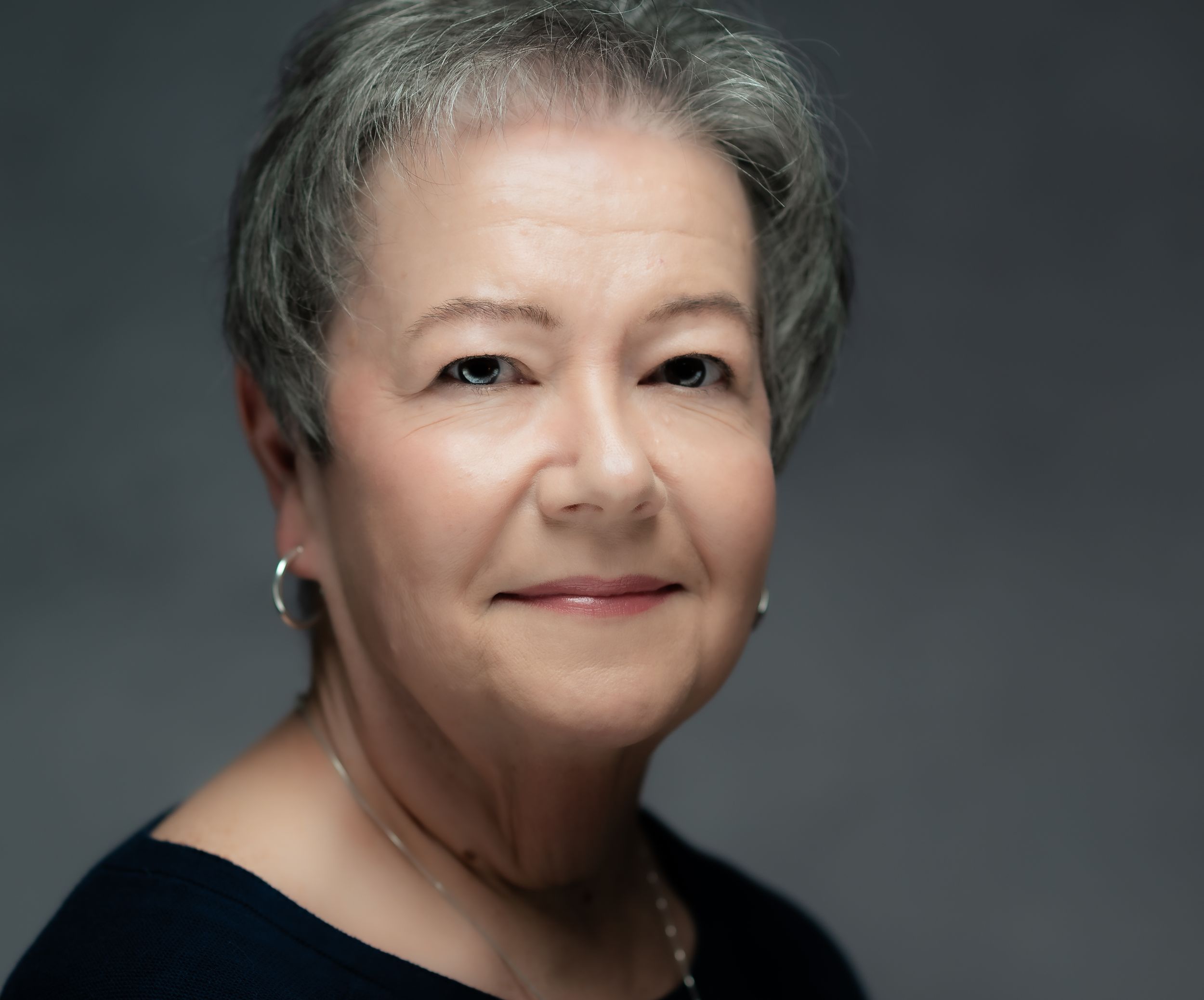 Professional headshot portrait of a mature woman with short gray hair wearing a black top against a dark gray background.