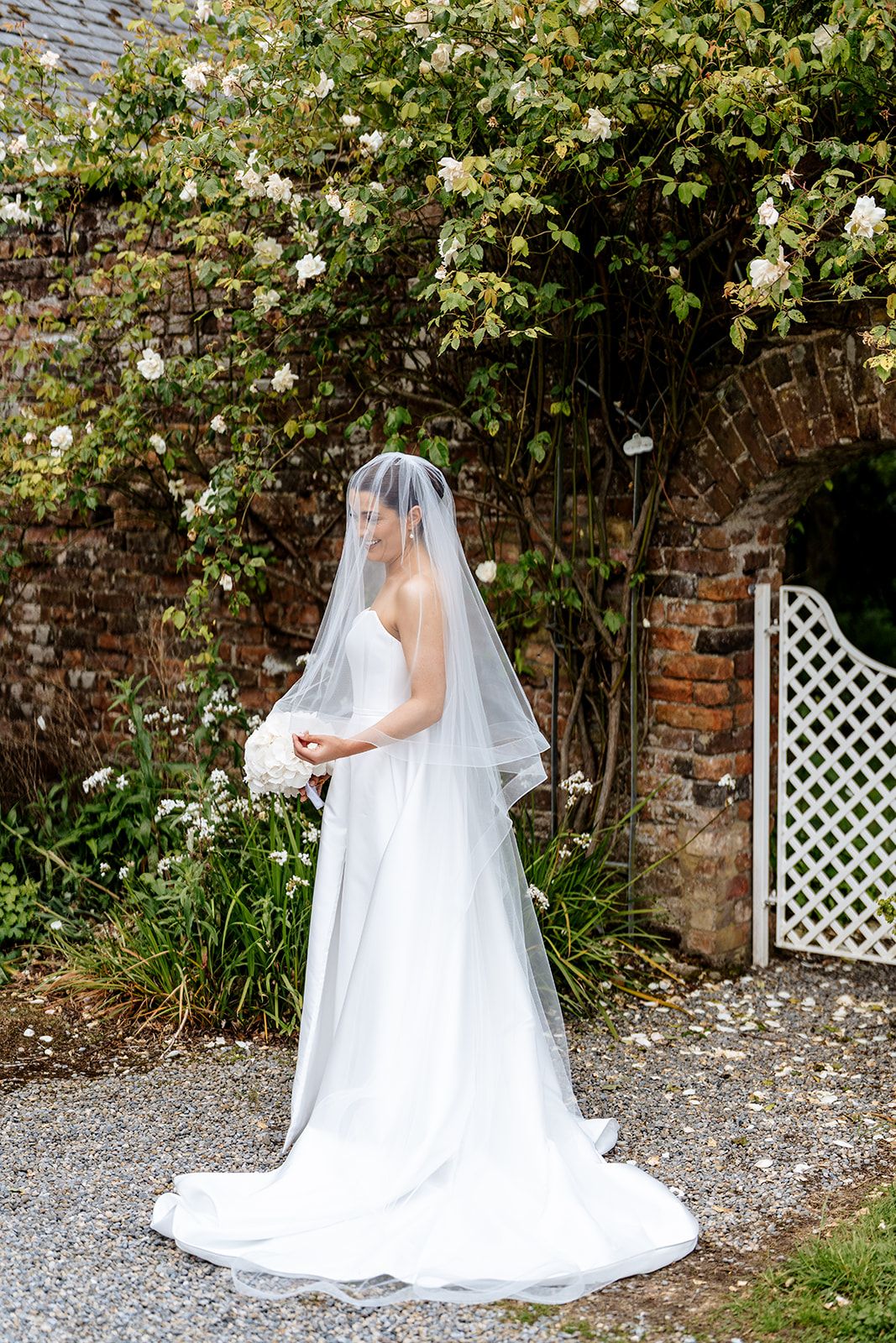 A bridal figure in a flowing white gown and long veil stands against a garden wall with climbing roses.