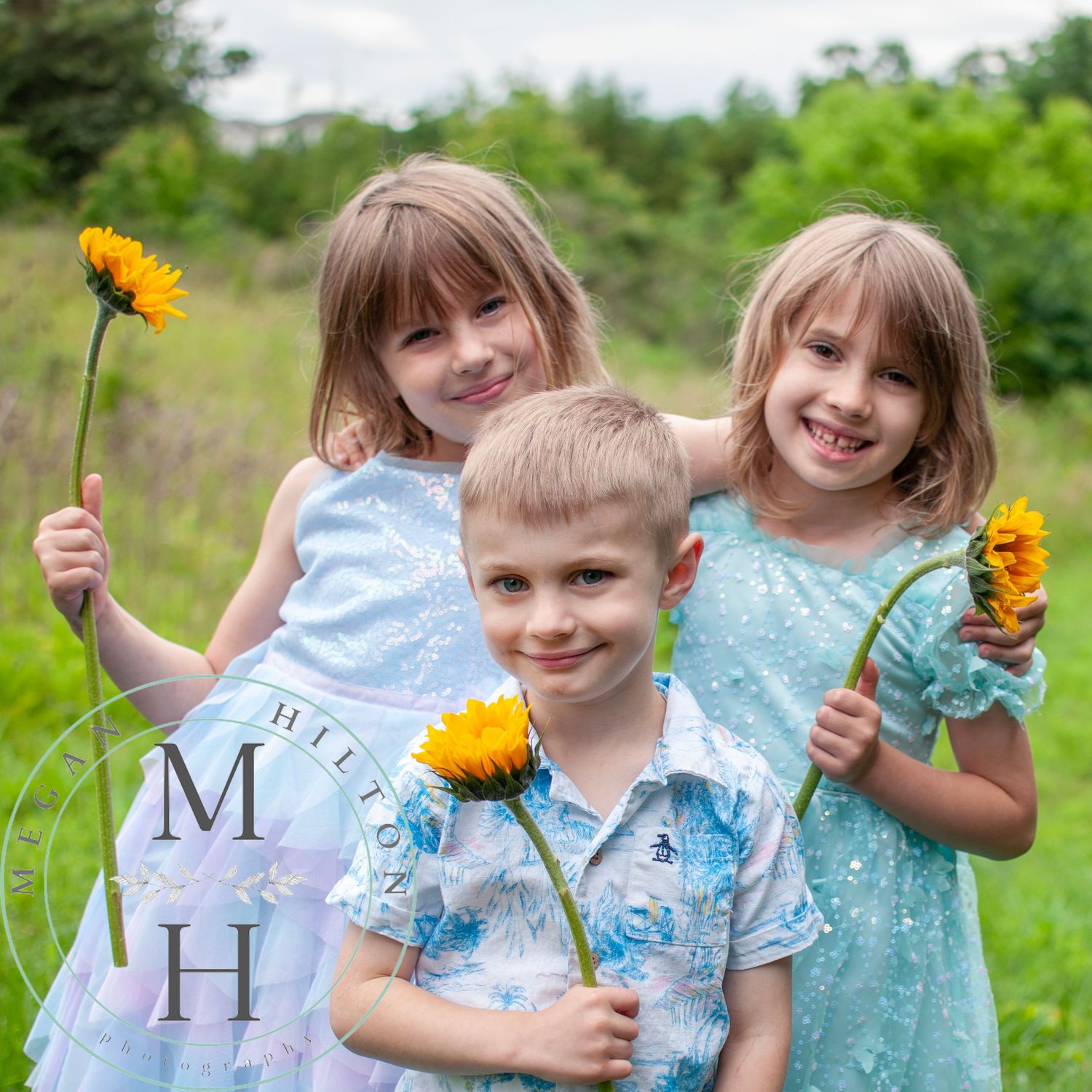 Children in blue and teal dresses hold bright yellow sunflowers flowers while playing outdoors in a grassy field.
