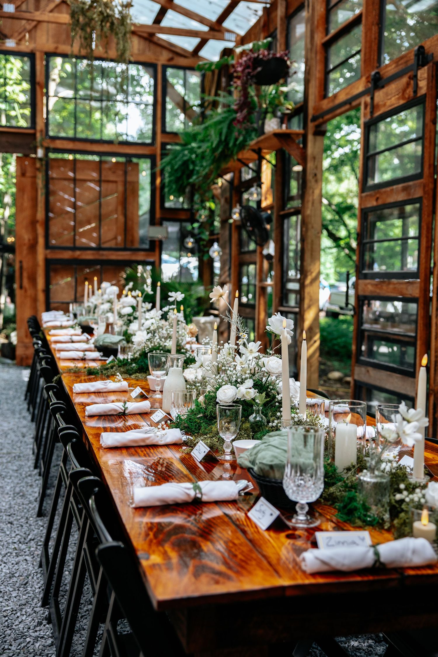 Elegant wedding table setting in a rustic greenhouse with wooden tables, white floral centerpieces and natural light.