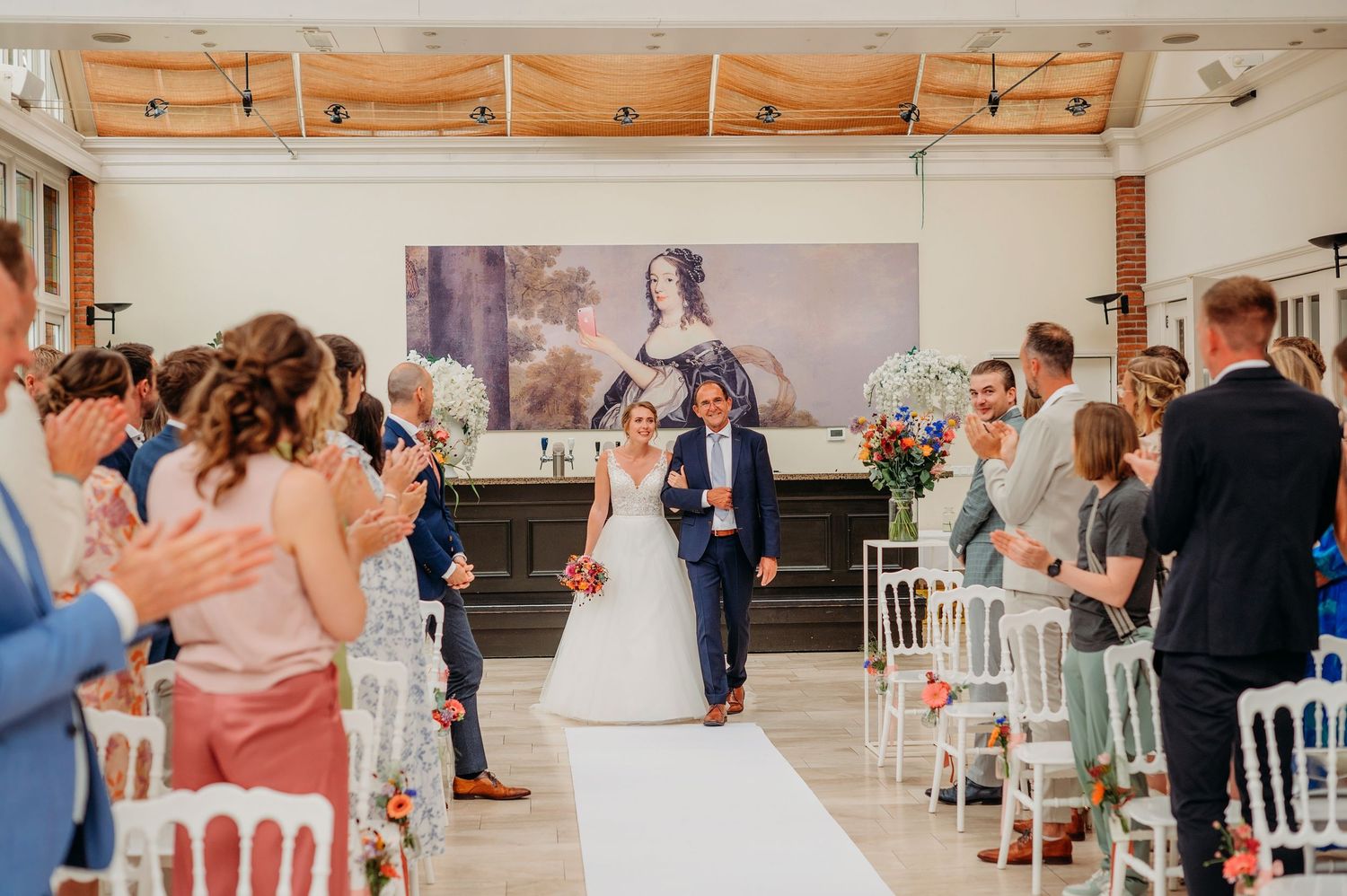 Wedding ceremony guests taking photos of couple walking down white aisle runner in bright indoor venue.