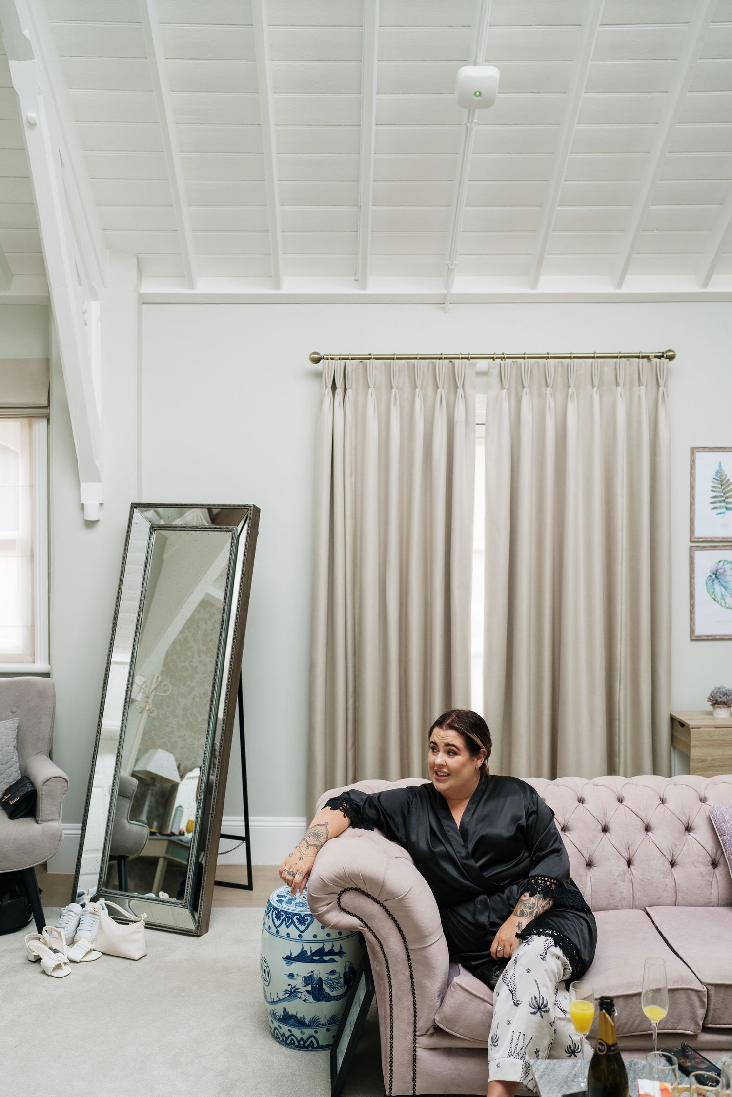 Interior room with white walls, large mirror, beige curtains and pink tufted sofa with someone relaxing in casual attire.