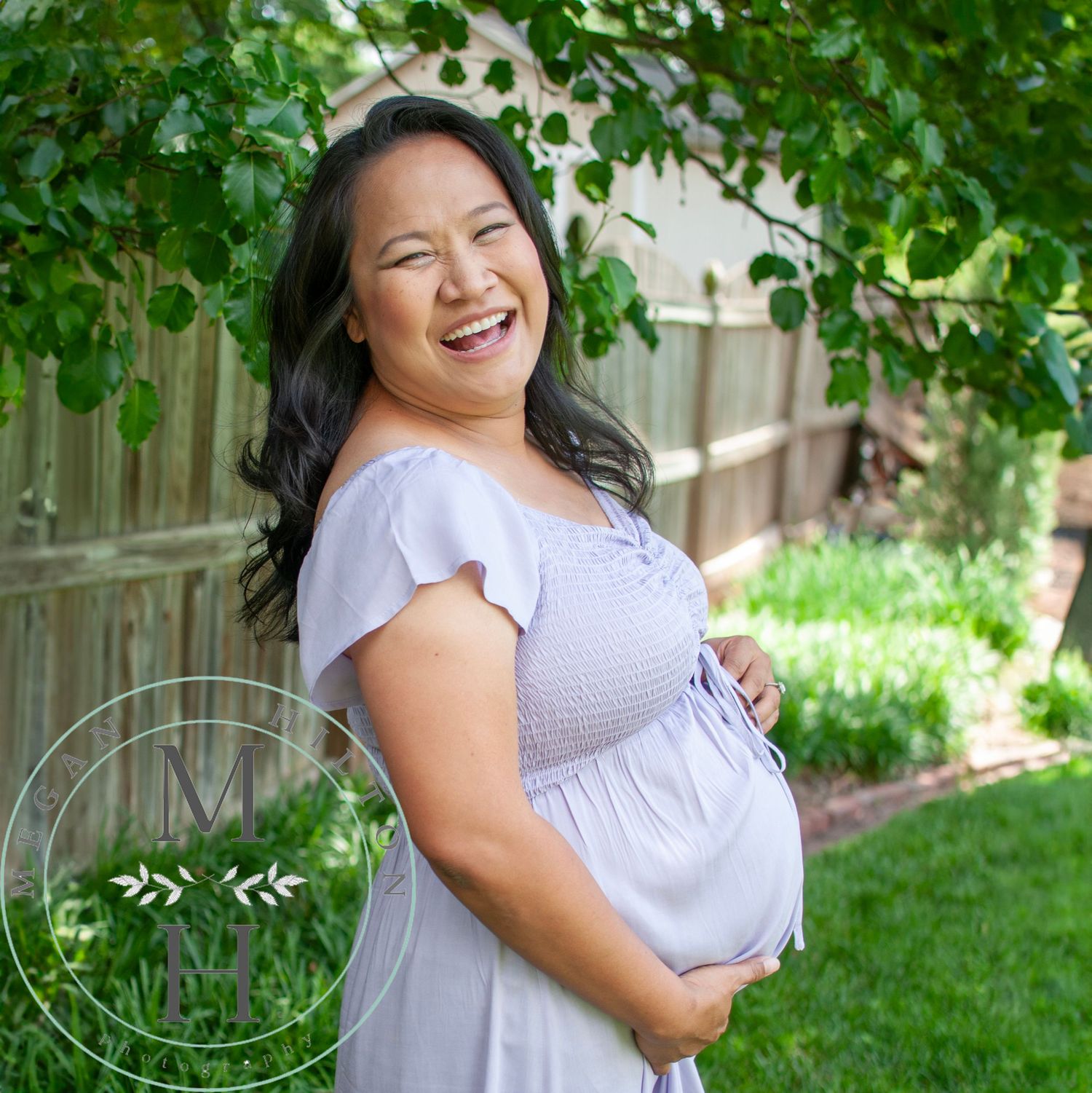 Smiling mama in a light purple dress poses for maternity photo outdoors with green foliage in the background.