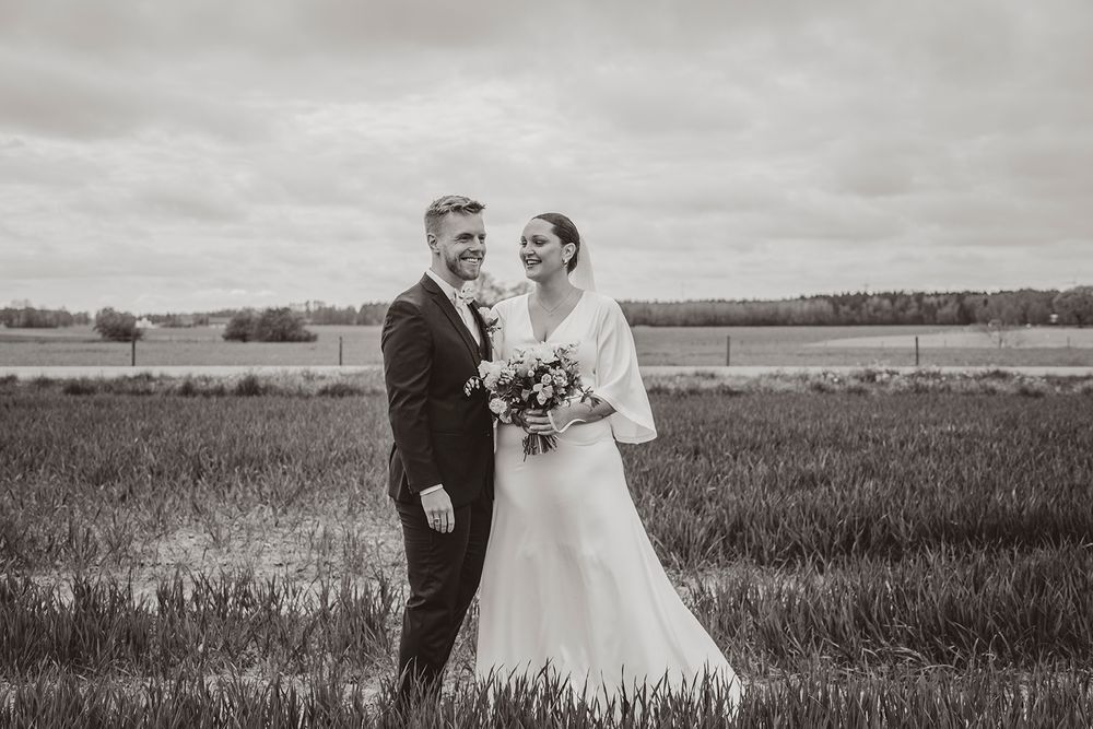 A couple stands together in a field on their wedding day under cloudy skies with open landscape in the background.