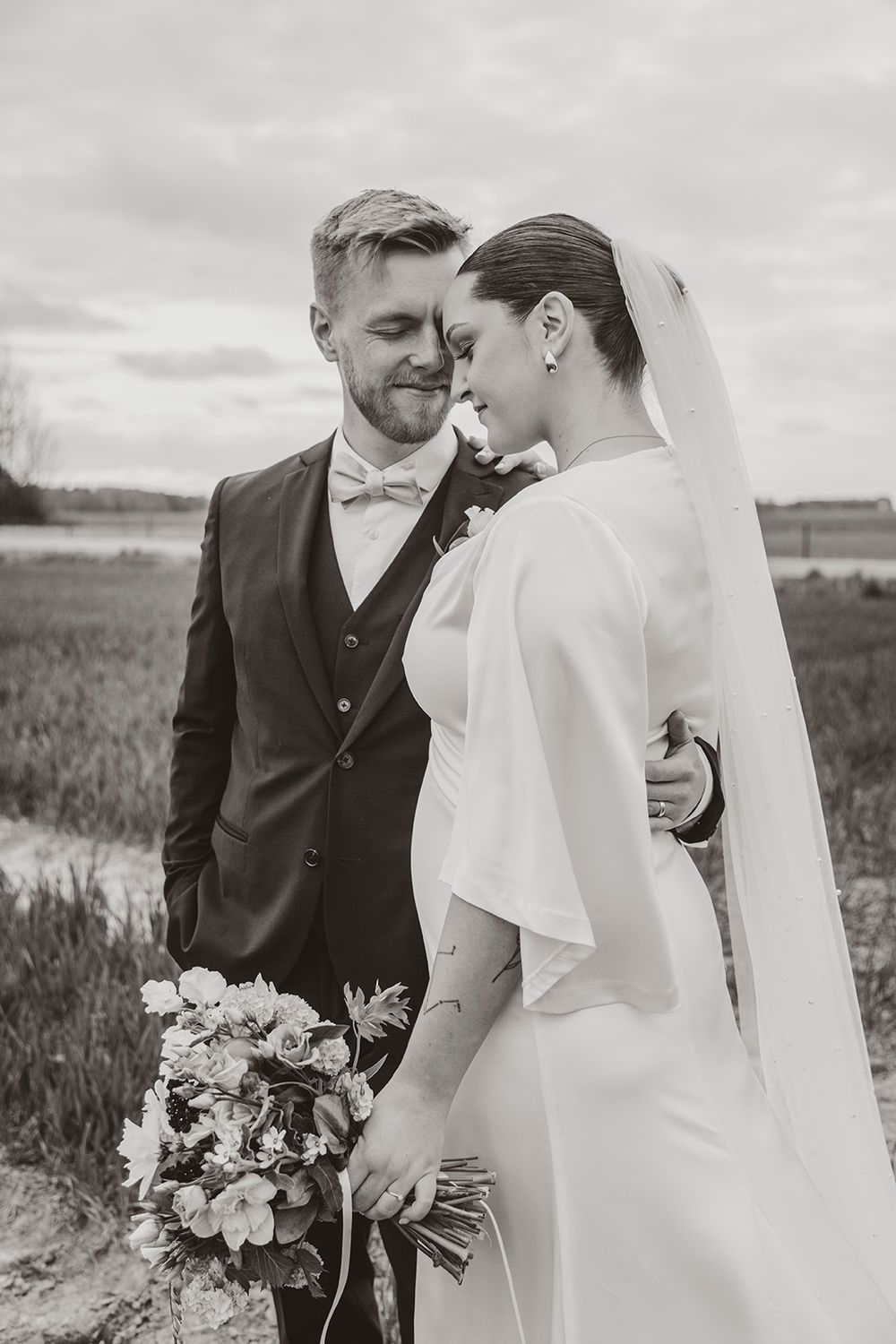 Black and white wedding photo showing a loving couple embracing in a field with the bride holding a floral bouquet.