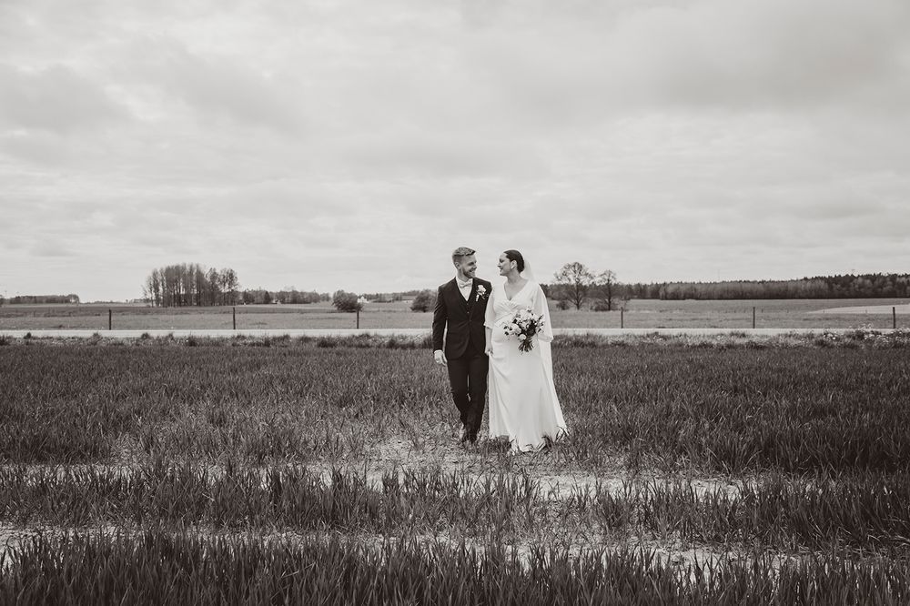 A vintage black and white wedding photograph showing a couple standing together in an open field with a lake in the background.