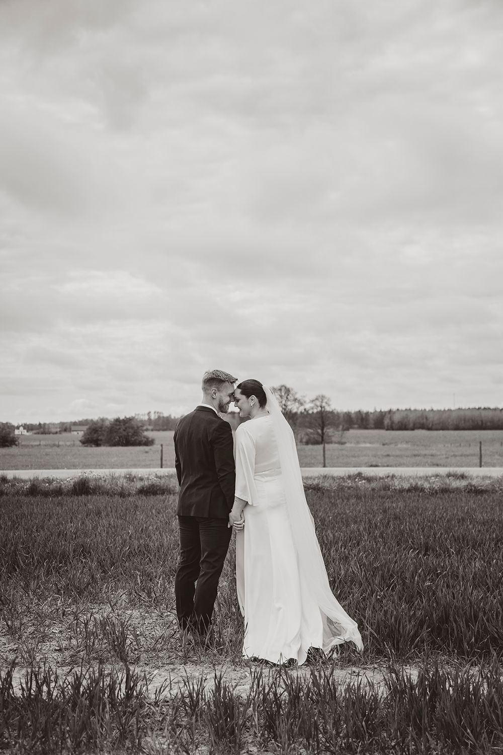 A black and white wedding photo shows a couple embracing in a rural field under moody overcast skies.