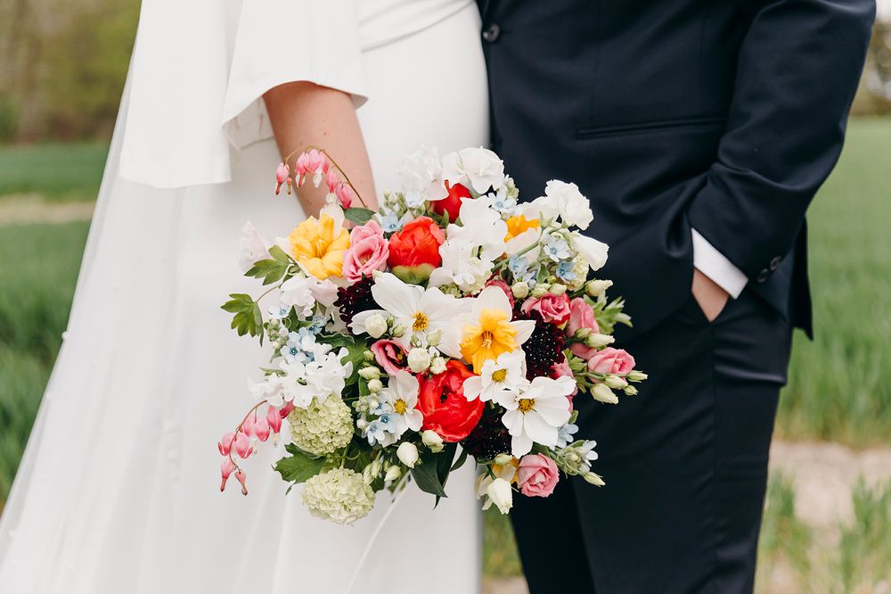 A vibrant wedding bouquet with red, orange, pink and white flowers held between a bride in white and groom in black formal wear.