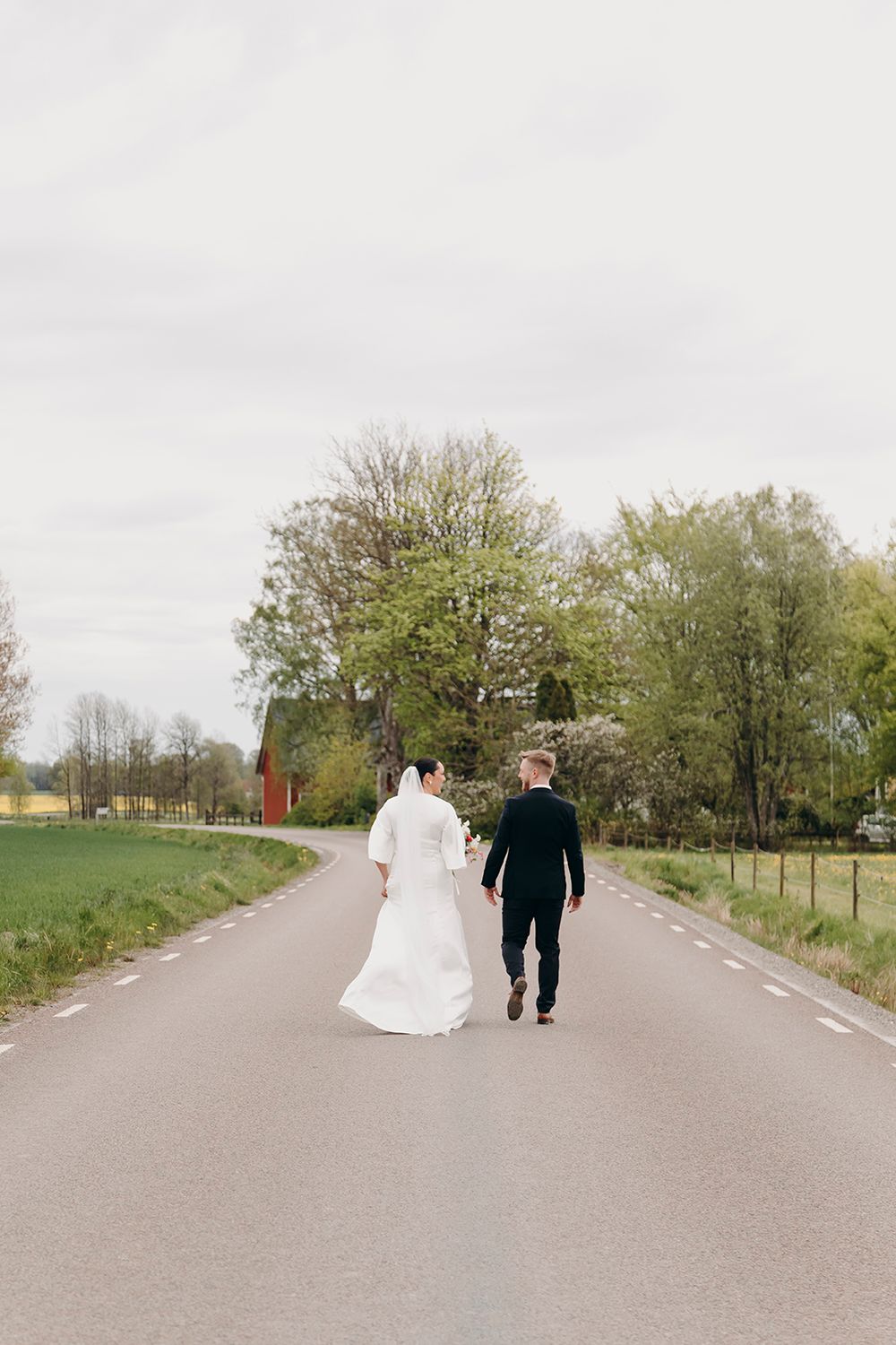 A couple walking away from the camera on a rural road, with the person in white wearing a wedding dress.