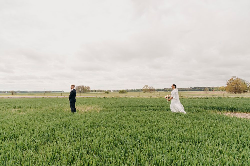 Two people stand apart in a green wheat field on an overcast day with farmland in the background.