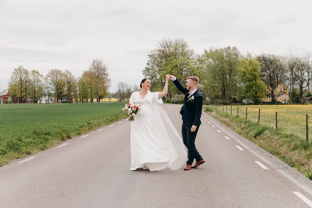 A couple in wedding attire dances joyfully on an empty country road lined with spring trees.