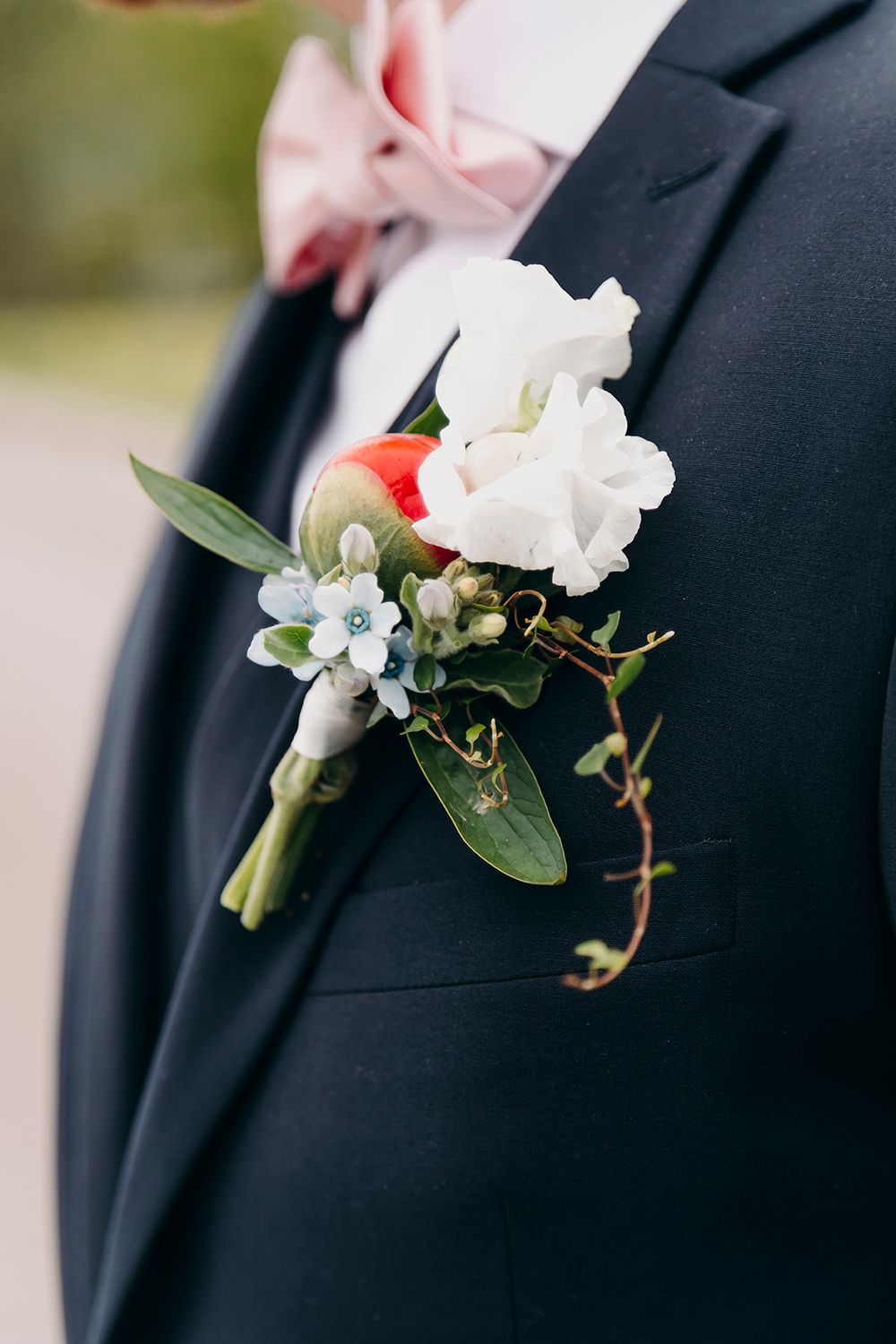 A white floral boutonniere with small blue accents pinned to a black suit lapel at a formal wedding.