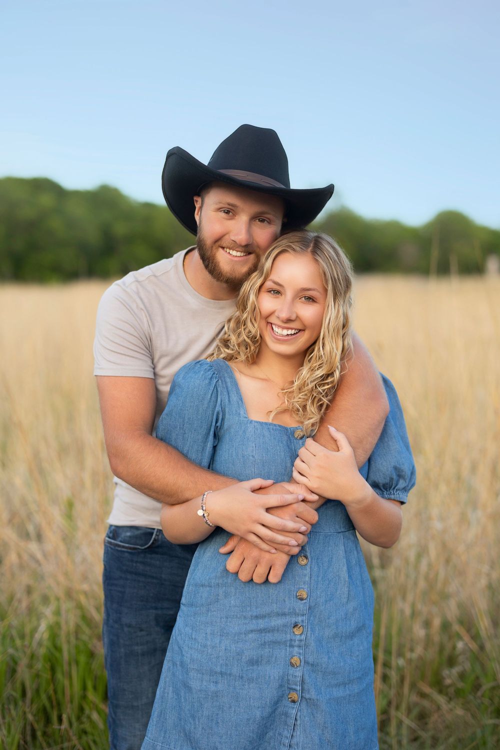 A couple embraces in a sunny field of tall grass, with the person in a cowboy hat and gray shirt hugging their partner in denim.