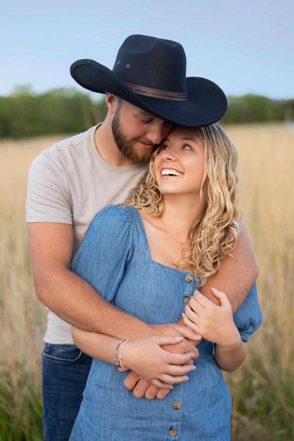 Romantic couple embracing in a wheat field at sunset, with her in denim dress and him wearing a cowboy hat.