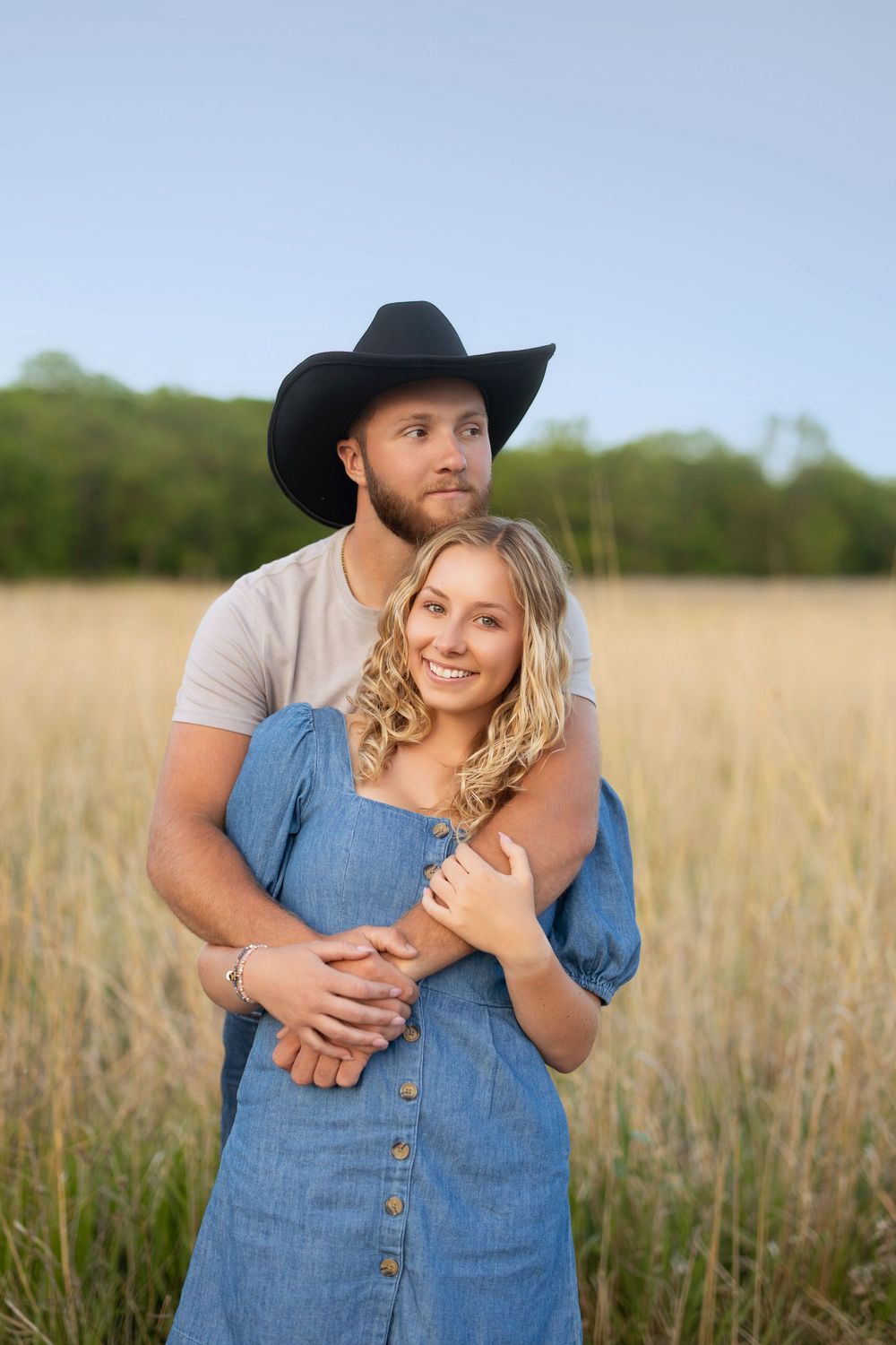 A romantic couple embraces in a golden wheat field at sunset, with the person wearing a black cowboy hat and gray shirt.