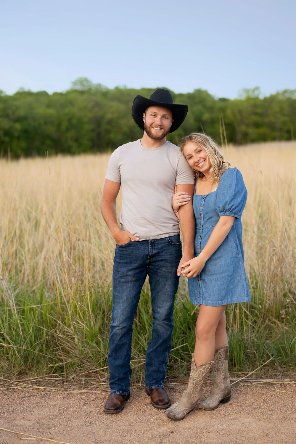 Couple standing together in tall grass field wearing country western attire with cowboy boots and hat.