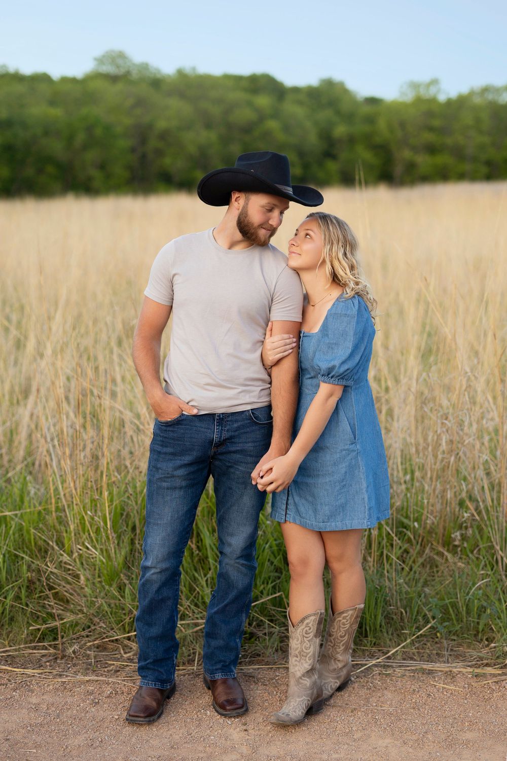 A couple in country western attire embrace in a field of tall grass during golden hour.