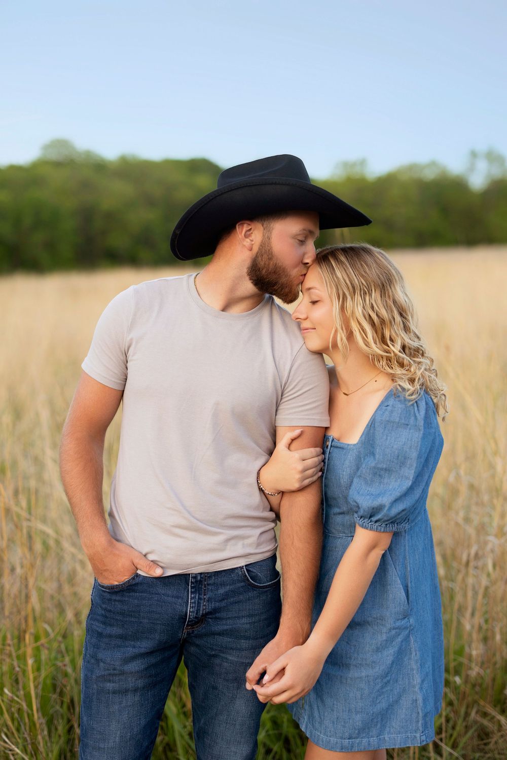 A romantic couple embraces in a golden wheat field at sunset, wearing denim and a cowboy hat.