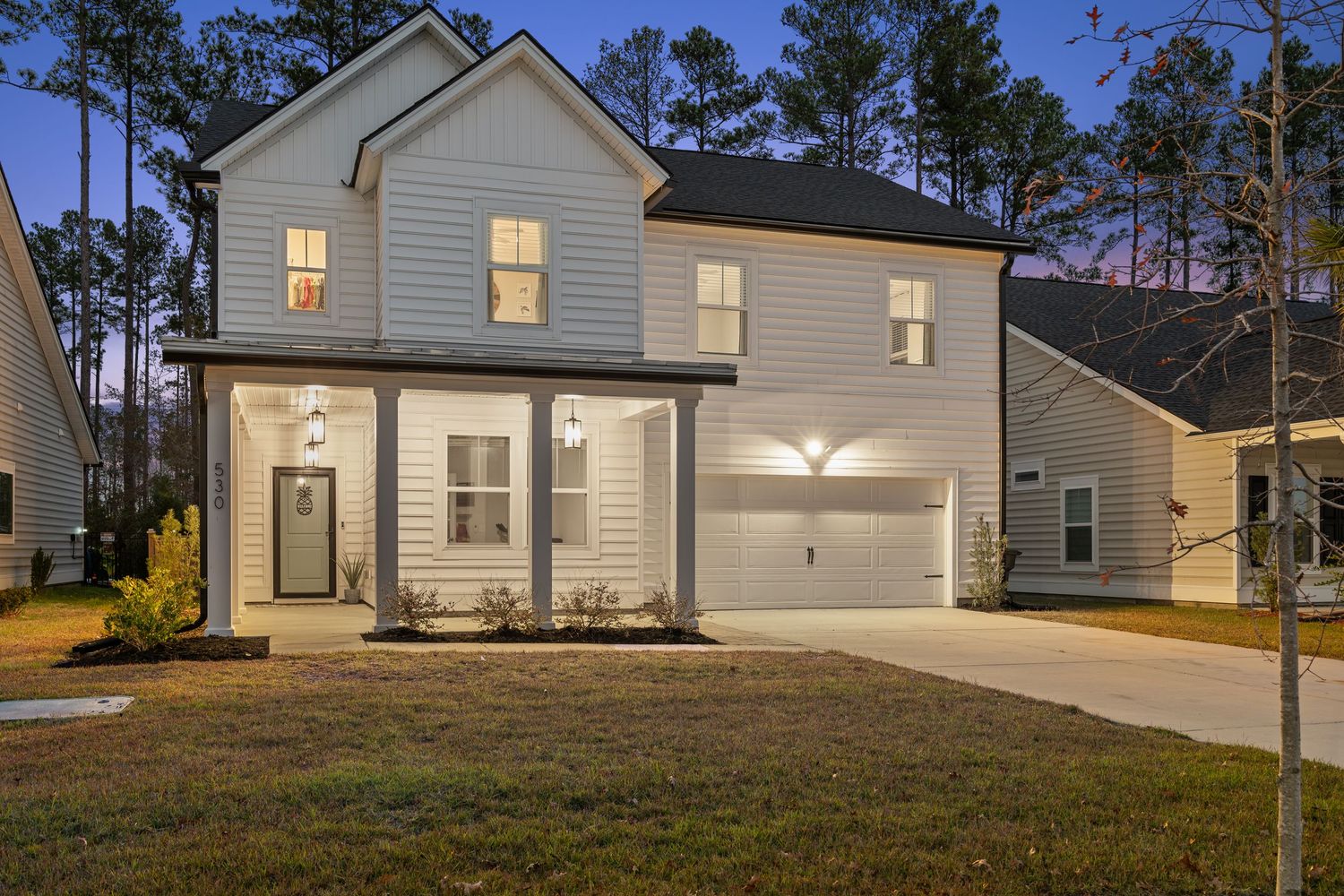 Modern two-story farmhouse with white siding and attached garage illuminated by warm light at dusk.