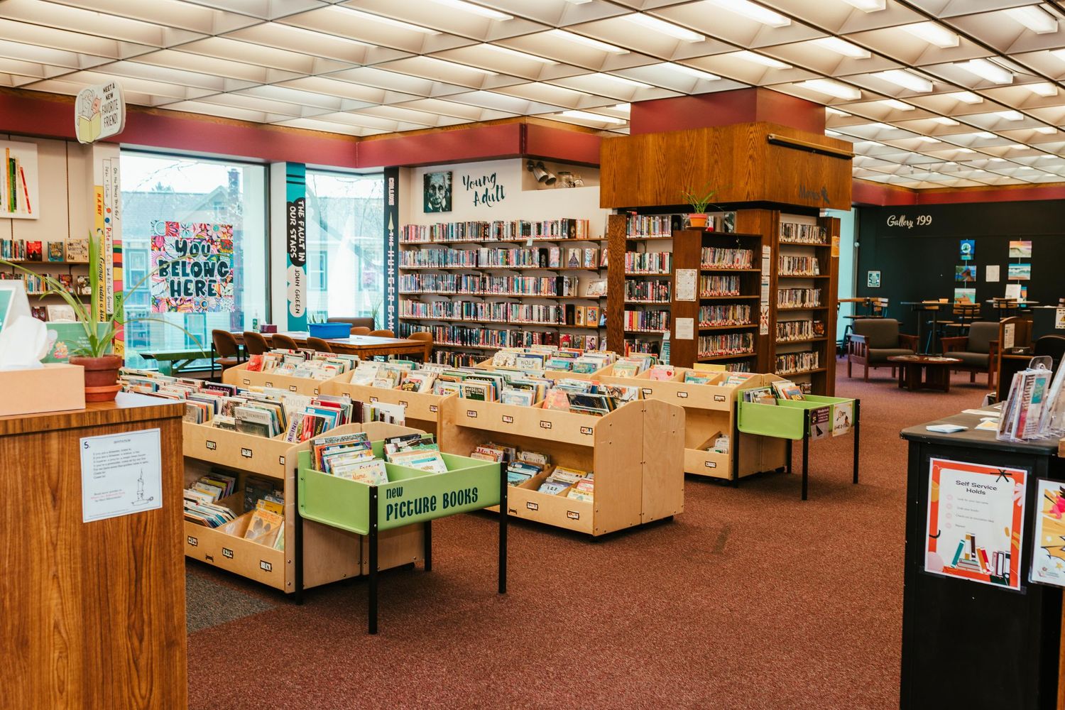 Modern Myerstown library interior with wooden shelves, book displays and green storage bins on brown carpeting under a grid ceiling.