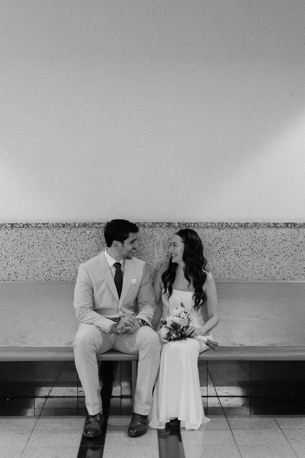 A newly married couple sits together on a bench sharing an intimate moment in black and white photography.