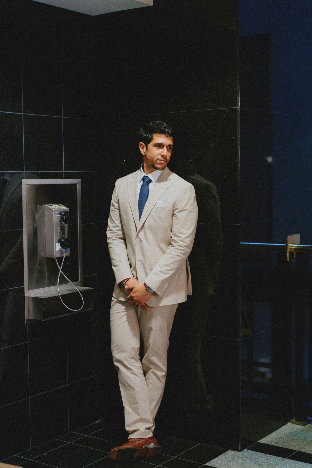 A person in a beige suit and blue tie stands against dark tiled walls near a payphone.