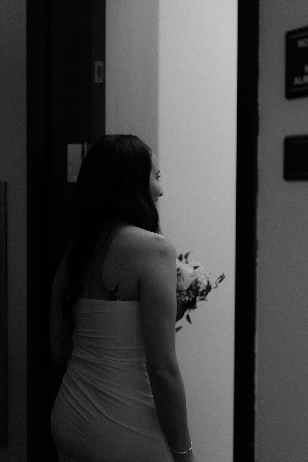 Black and white portrait of a person in a white dress seen from behind standing in a dimly lit hallway.