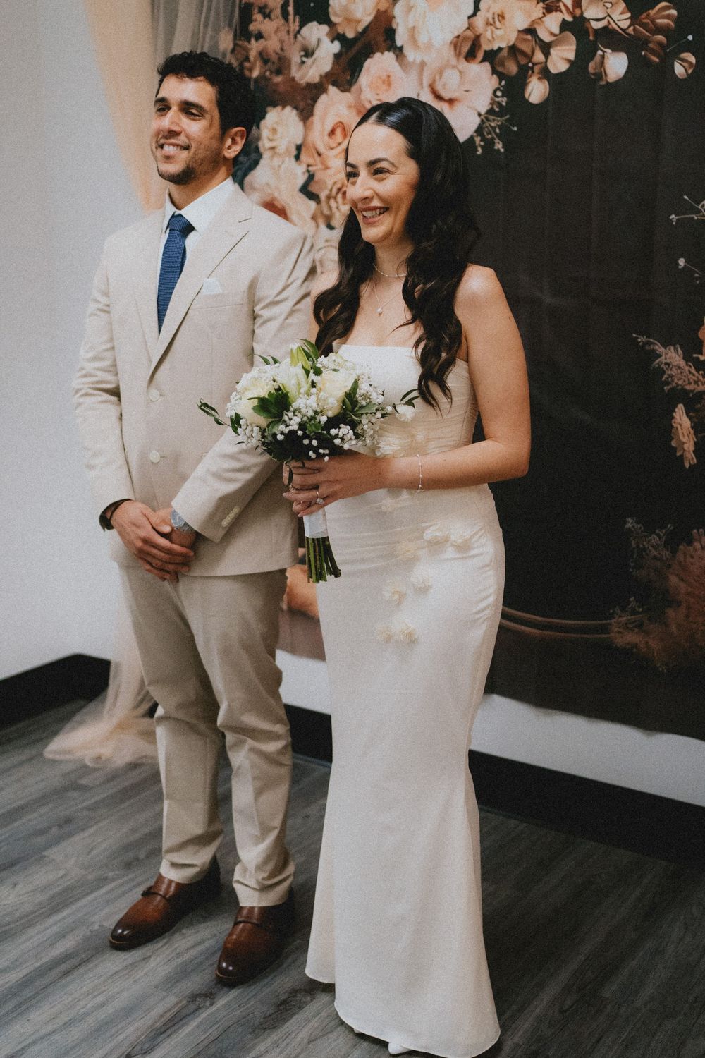 Newly married couple share a candid laugh during wedding portraits against a floral backdrop on gray wood floors.