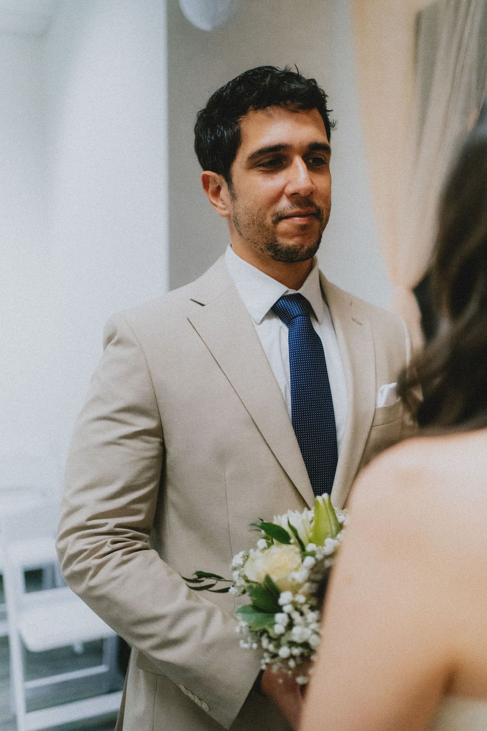 Groom in a light beige suit and blue tie holds a small white floral bouquet during a wedding ceremony.