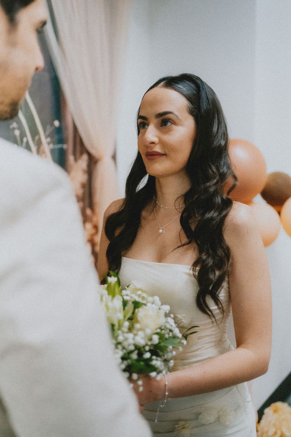 A couple shares an intimate moment at their wedding ceremony with orange balloons and white floral bouquet in soft lighting.