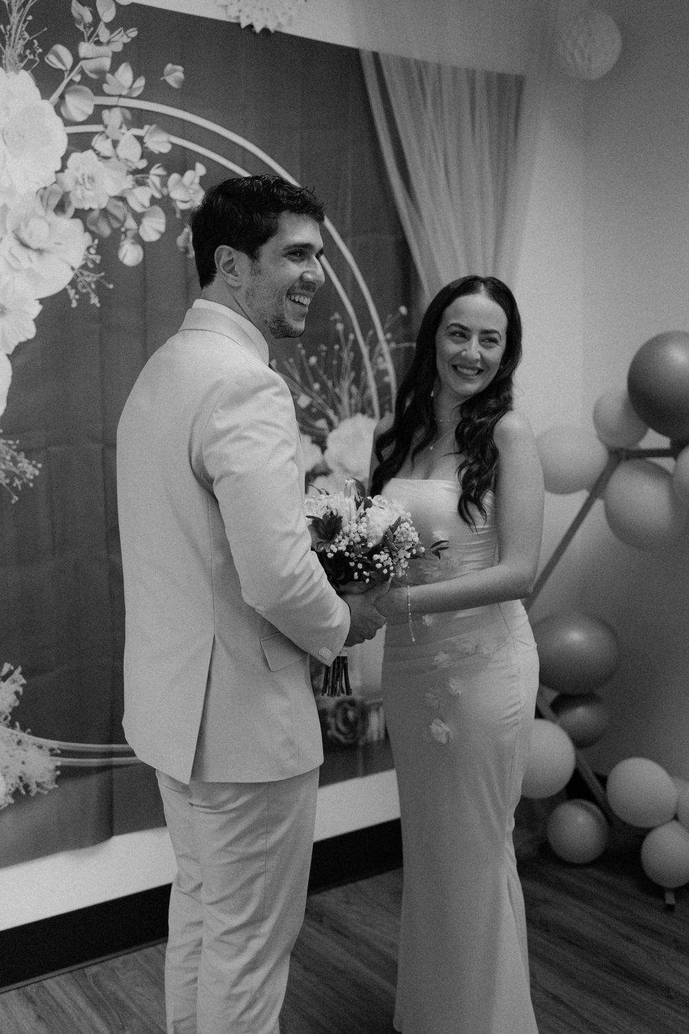 A newlywed couple smiles while holding a bouquet in front of a floral backdrop with white balloons.