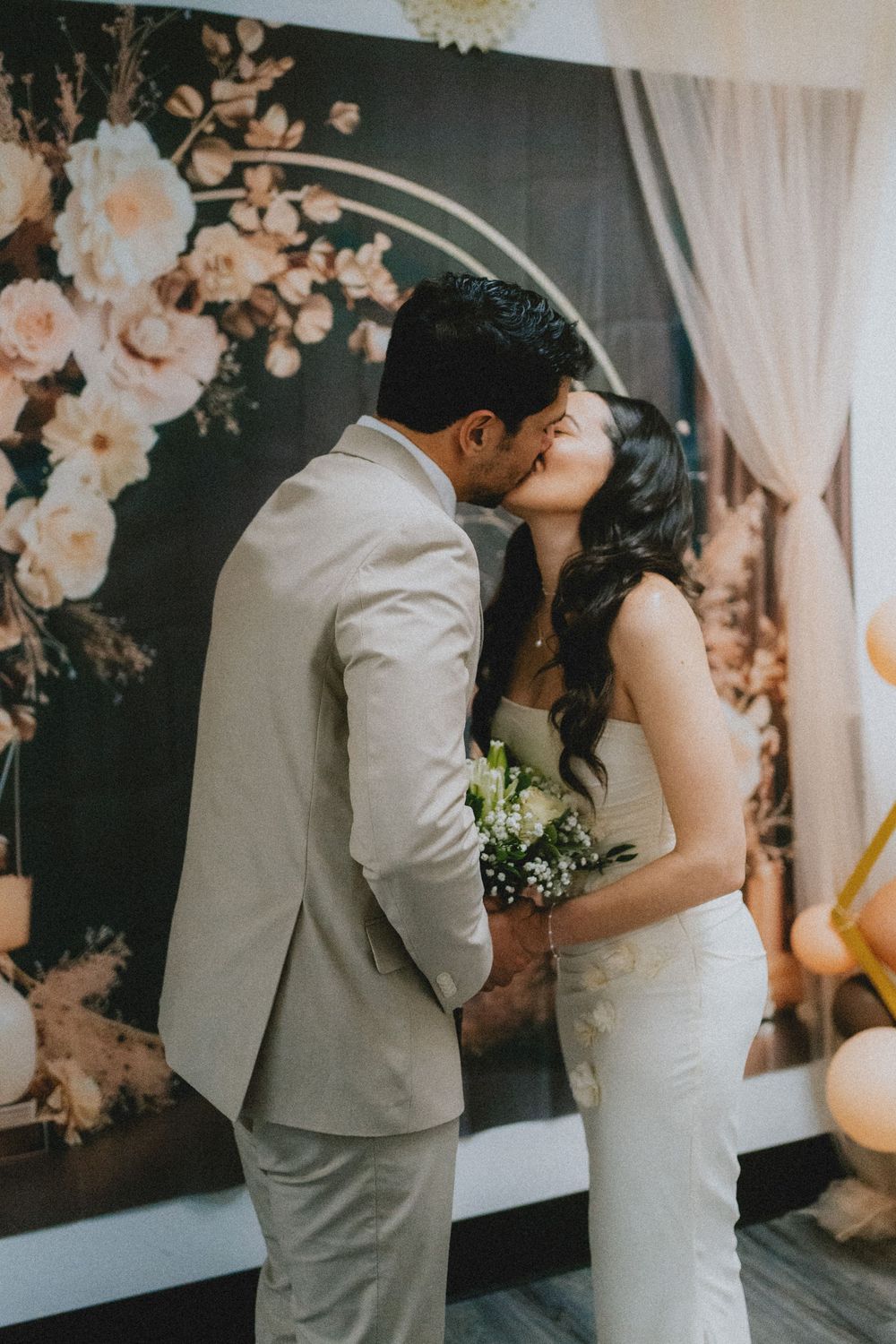 A romantic wedding kiss captured in front of a floral arch with peach roses and white curtains.
