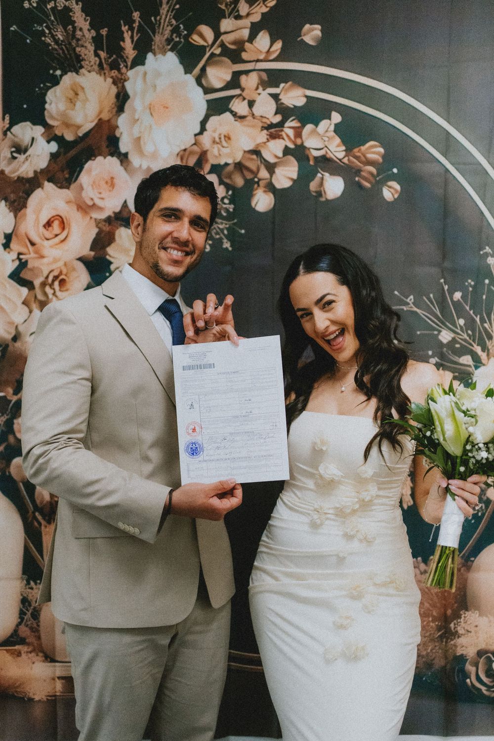 A newlywed couple shows off their marriage certificate in front of a floral backdrop at their wedding ceremony.