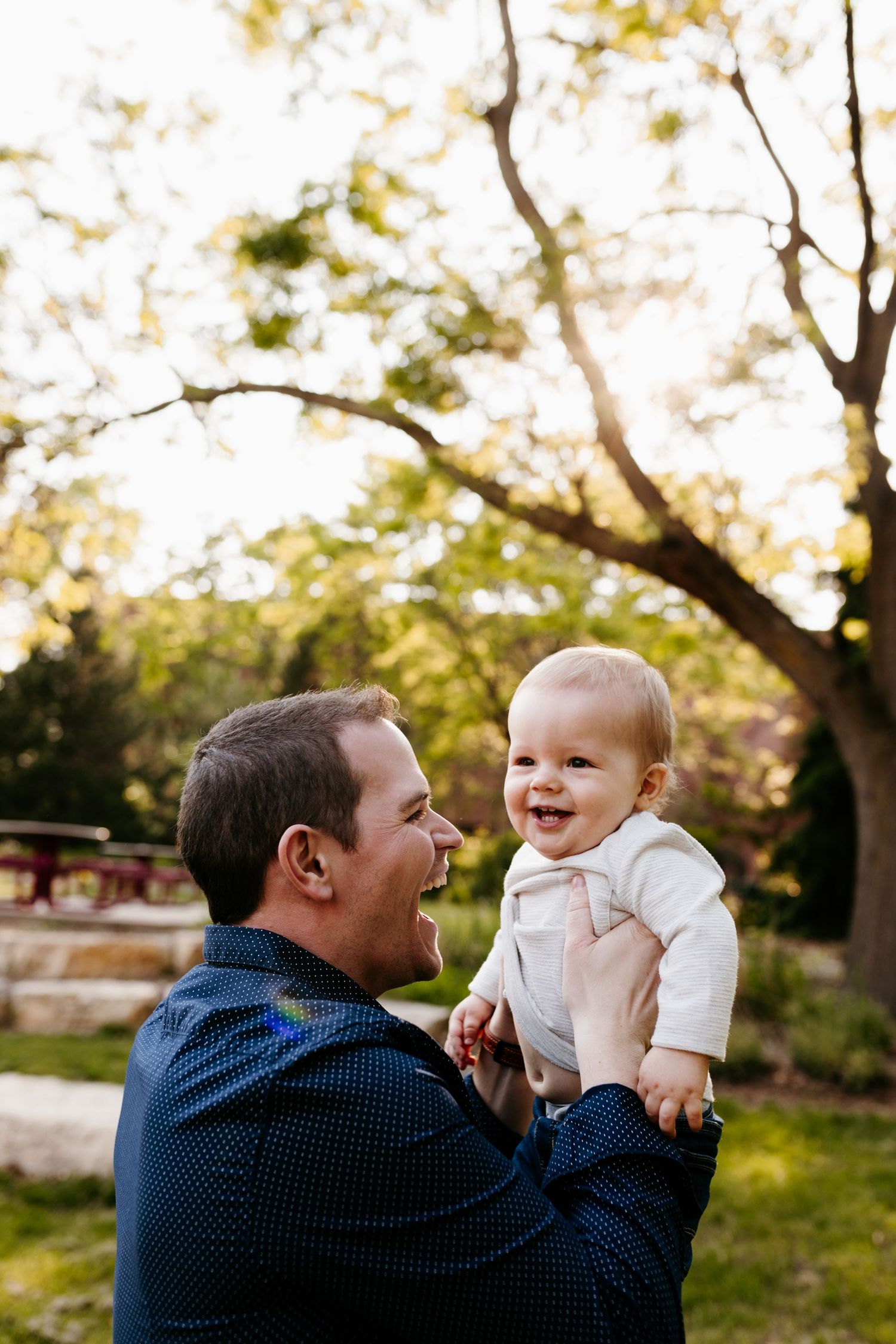 A baby in white clothing laughs joyfully while being held up outdoors during golden hour with trees in the background.